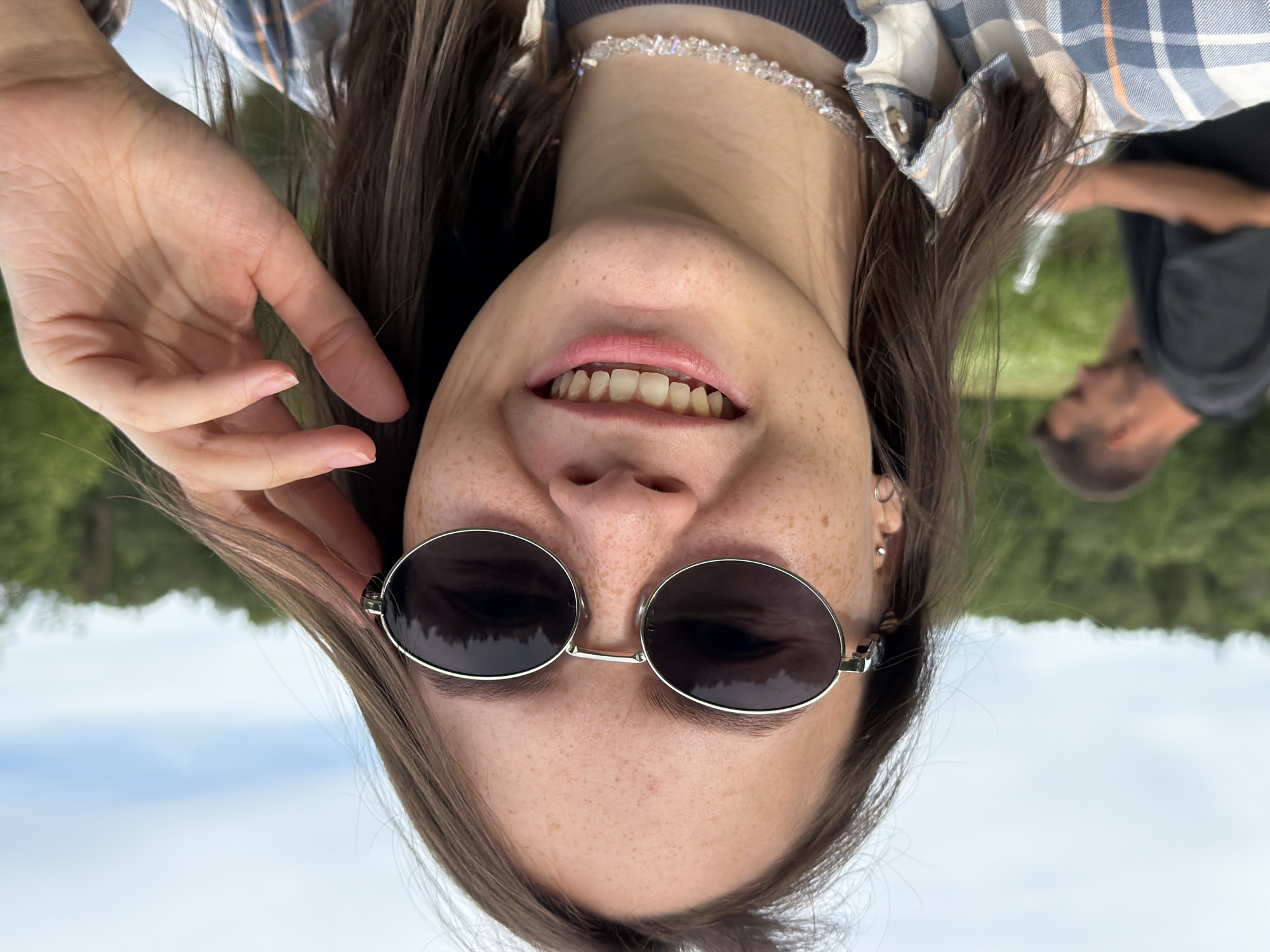 The image is a close-up selfie of a young woman with brown hair and fair skin, wearing sunglasses and a plaid shirt. She has freckles on her face and is smiling with her teeth showing. She is wearing a beaded necklace. Her right hand is raised, touching her hair near her temple. In the background, slightly out of focus, is a man wearing a dark t-shirt. The setting appears to be outdoors with a blurred green, tree-filled landscape and a pale sky with clouds. The location is the city of Garevaya, Russia.