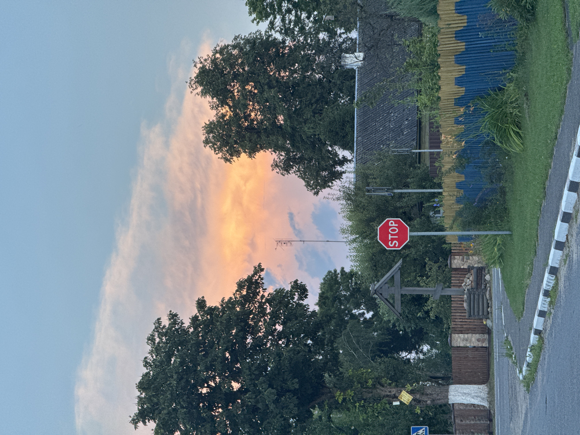 The image captures a rural roadside scene in the Neman Rural Council, Belarus, at dusk. The sky is a blend of soft blues, pinks, and oranges, indicating the sun has recently set. In the foreground, a paved road curves to the left, bordered by a curb with black and white markings. A green grassy verge runs alongside the road. To the right, a vibrantly painted fence, featuring a wavy pattern of blue and yellow, delineates the property line. Behind the fence, dense greenery and the roof of a building are visible.
Dominating the mid-ground is a large, mature tree on the left side of the road, its trunk partially whitewashed. Next to it stands a wooden roadside cross. A red octagonal stop sign is prominently displayed on a pole in the center of the frame, facing the road. Further back, behind the fence and trees, a television antenna stands tall. The overall impression is of a tranquil, rural setting bathed in the soft light of twilight. No people are visible in the image.