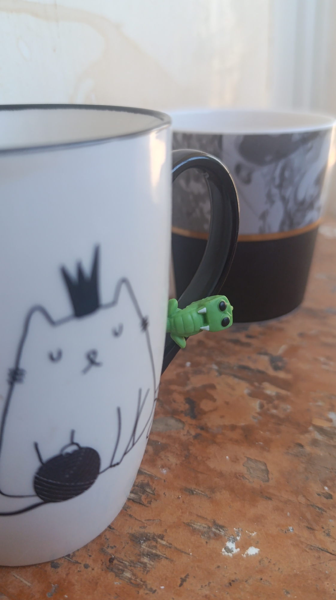 A close-up, eye-level shot shows two mugs on a wooden surface. The mug in the foreground, on the left, is white and features a black outline drawing of a cat wearing a crown. A small, green, plastic alligator figurine is attached to the handle of this mug. The mug in the background is black with a gray marbled pattern. The surface the mugs are on appears to be a wooden table, with visible grain and some white specks. The background is blurred, showing a wall with water stains and diffused light. The image was taken in an outdoor setting, possibly a patio or balcony, based on the natural lighting and the wooden surface.