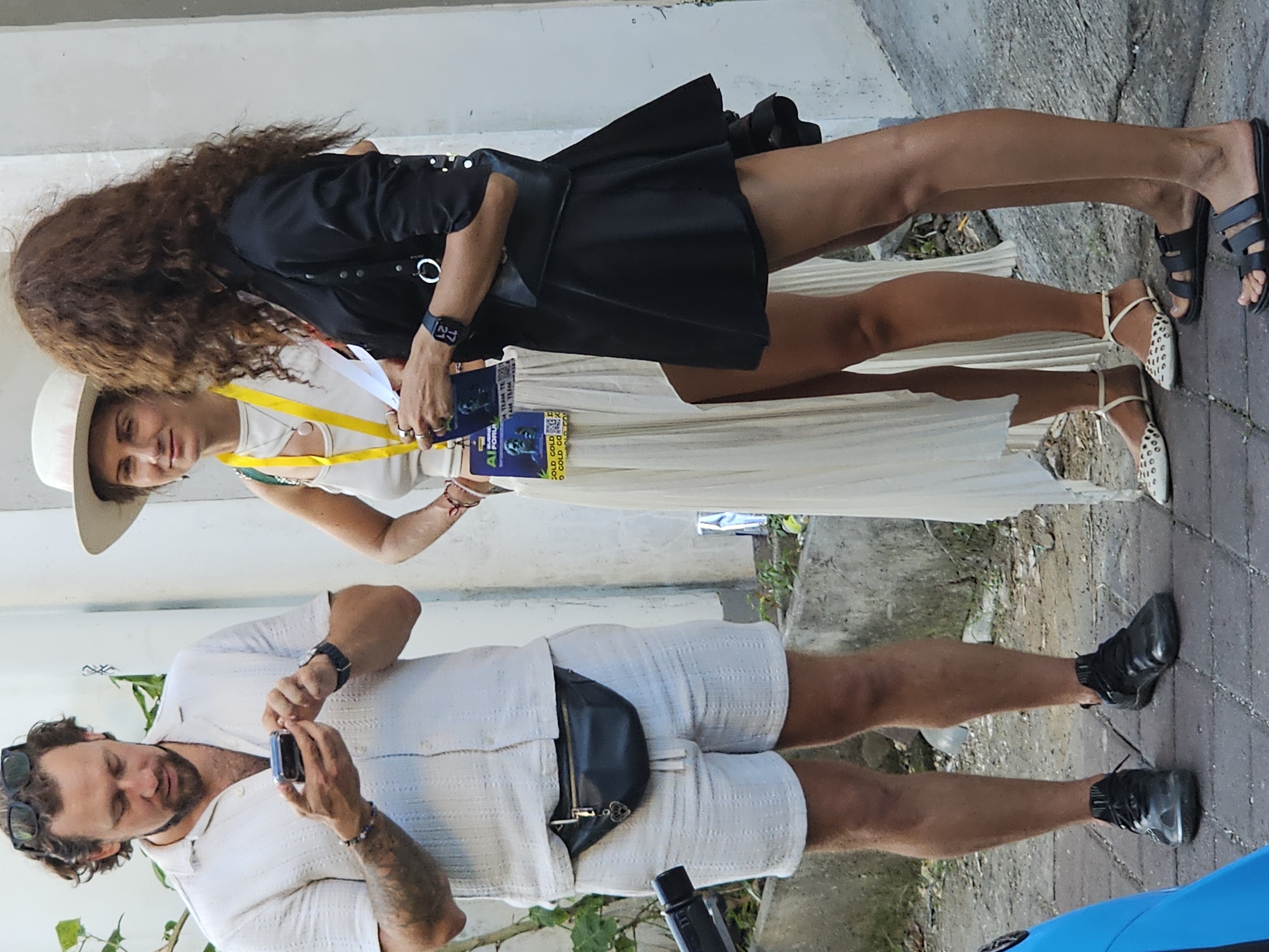 A man and two women stand outdoors in North Kuta, Indonesia. The man, wearing a white shirt and shorts with a black fanny pack, is looking at a smartphone in his hands. One woman is wearing a white dress and a straw hat, and the other is in a black dress and sandals. Both women have passes hanging around their necks. They are standing near a white wall with foliage nearby.