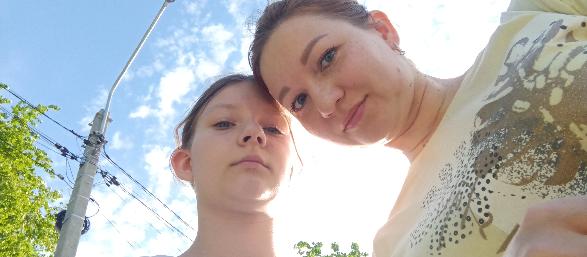 Two people, a younger and older female, are positioned close together in an outdoor setting. The camera angle is low, looking upwards. The older female has light skin, her hair is pulled back, and she is wearing earrings and a patterned top. The younger female has lighter skin and her hair is also pulled back. Behind the subjects is a sky with sparse clouds. To the left is a concrete utility pole with visible wiring, and green foliage is visible near the bottom and left of the frame. The location is a city, most likely unknown.