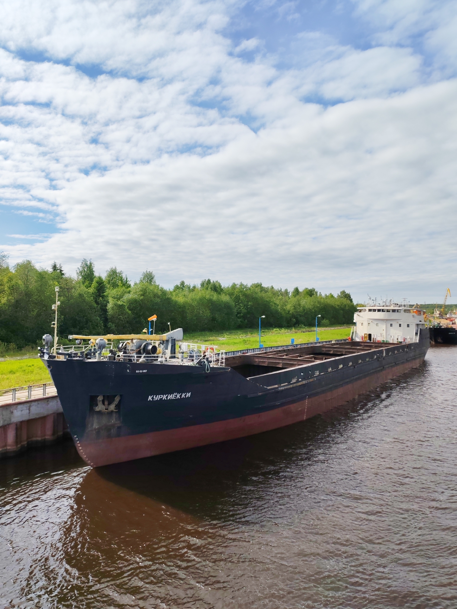 A barge named "Kypkuekku" sits on the water next to a dock with a green lawn behind it. The barge is dark blue on top and burgundy on the bottom. The barge is near a city but the location is Unknown.