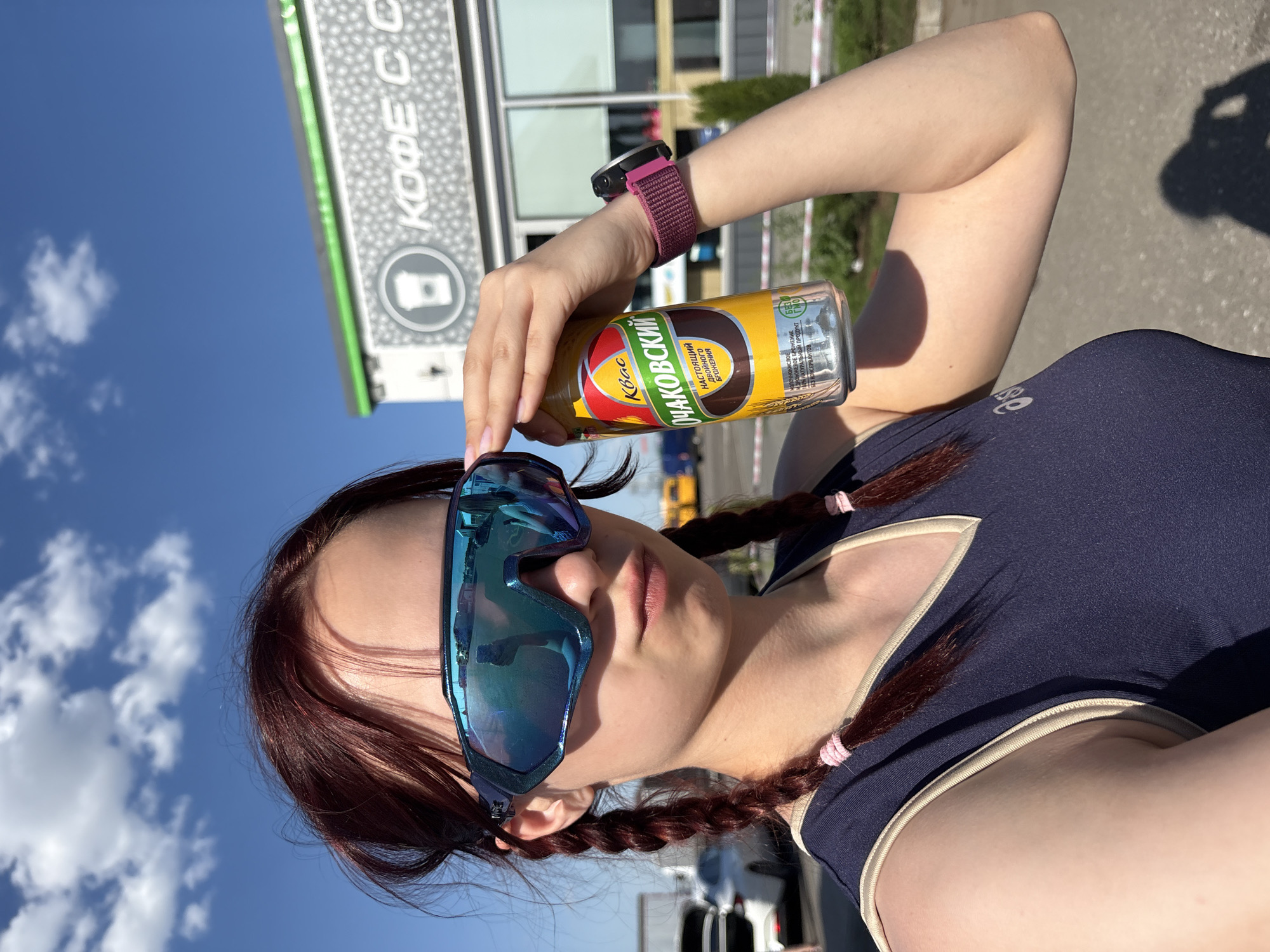 Here is a description of the image:
The photo depicts a young woman outdoors, holding a can of "Ochakovsky" kvass, a traditional Russian beverage made from fermented rye bread. She is wearing mirrored sunglasses and a dark blue top with tan trim. Her reddish-brown hair is styled in braids with pink ties. A smartwatch with a maroon band is visible on her left wrist.
The background features a building with signage in Russian. The text says "КОФЕ С С" which translates to "COFFEE C C". From the sign the location is most likely in the city of Kazan, Russia. The sky is blue with scattered clouds, suggesting a sunny day. A paved area is also visible, indicating she is likely in an urban setting.