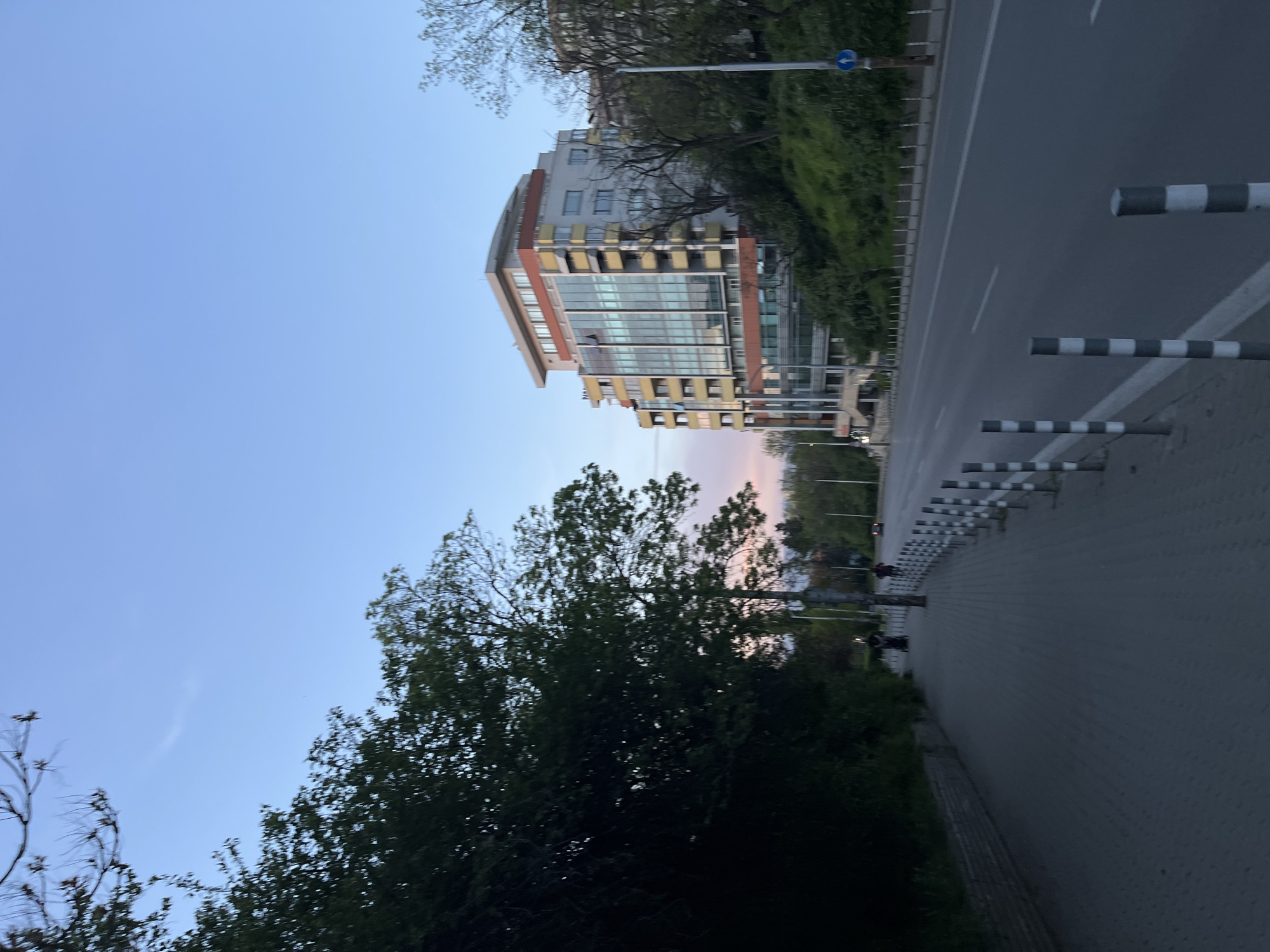 An urban street scene unfolds during dusk or early evening in Sofia, Bulgaria. The sky, a clear expanse of light blue, gradually fades into a soft, pale pinkish-orange hue on the horizon, indicating the setting sun.
On the right side of the image, a contemporary multi-story building dominates the view. It features extensive glass facades, a curved roofline, and numerous balconies, showcasing a blend of grey, brown, and yellow architectural elements. Behind this structure, lush green trees and bushes cover a hillside, with another building partially visible among the foliage further back.
Below the main building and running parallel to it is a paved road, characterized by its dark asphalt and a white dashed line indicating lanes. Adjacent to the road is a grey paved sidewalk or pathway. A series of black and white striped bollards line the edge of this pathway, acting as a barrier between the pedestrian area and the road. A blue circular traffic sign and a street light pole are visible near the road on the right.
On the left side of the frame, the silhouettes of dark green trees and dense foliage are prominent, obscuring the view beyond. The dimming light of the evening casts long shadows and deepens the colors of the vegetation.
The scene appears quiet, with no significant activity taking place. While faint figures might be discernible in the distance on the pavement, they are not clearly identifiable as people. There is no visible text on signs or buildings that can be clearly read. The overall impression is one of a peaceful urban landscape transitioning from day to night.