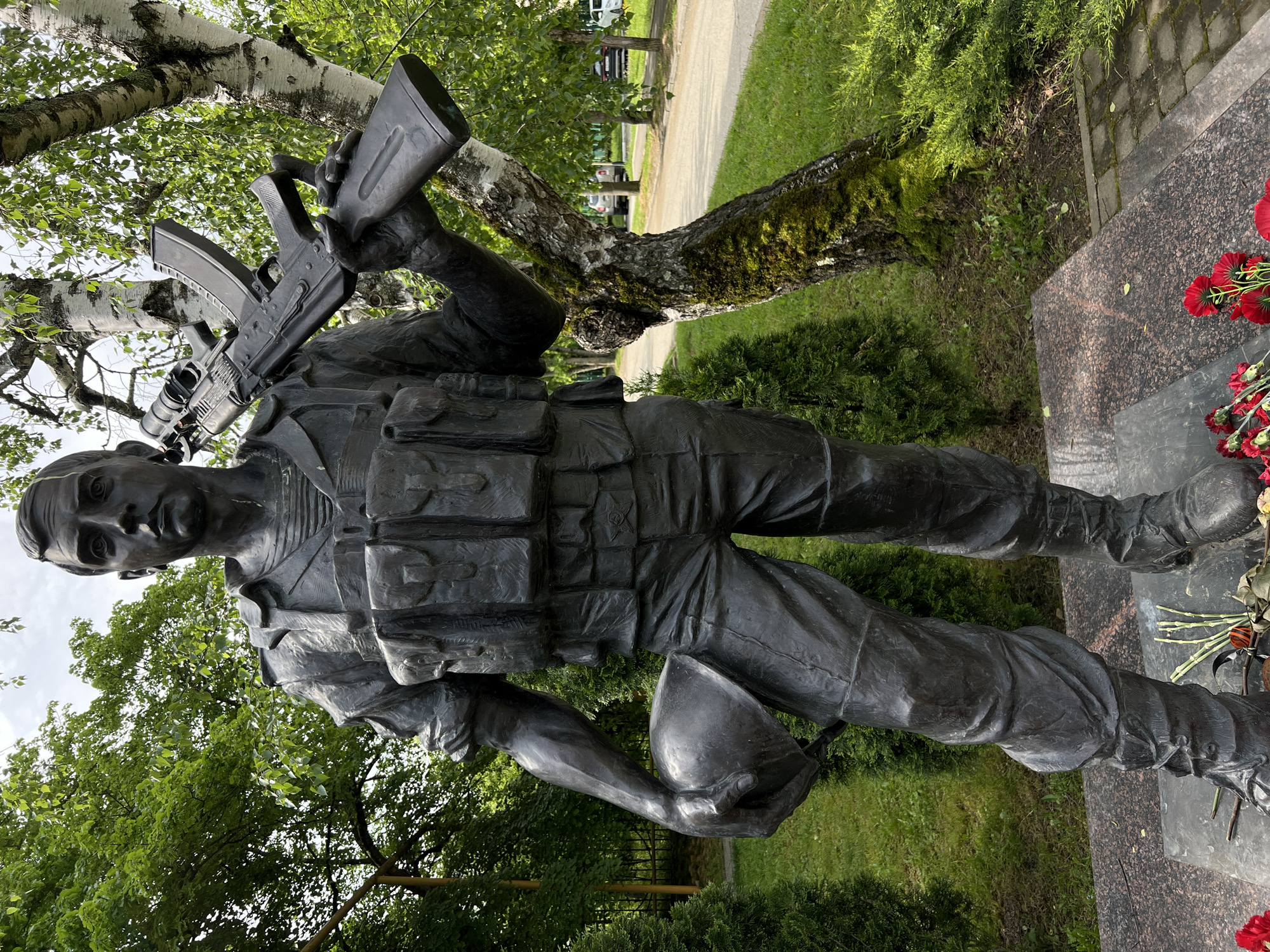 A statue of a soldier stands in Goryachiy Klyuch, Russia. The statue depicts a man in military uniform, holding a rifle over his shoulder and a helmet in his other hand. He is standing next to a tree and a stone platform with red flowers. The surrounding environment includes green grass, trees, and a paved pathway.