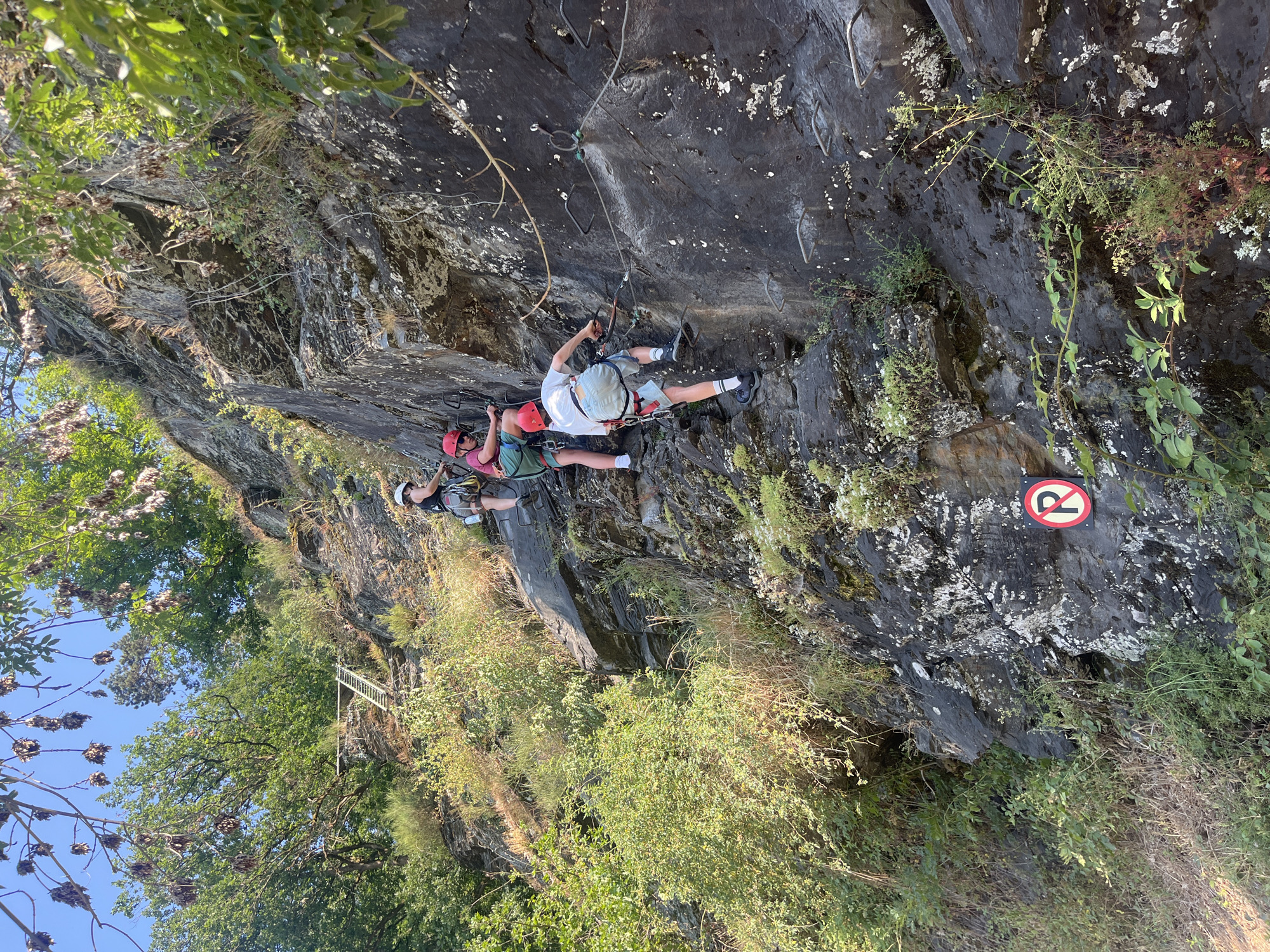 Three individuals are rock climbing on a steep, rocky cliff face. They are all wearing helmets and safety harnesses, and appear to be following a via ferrata route, indicated by the cables and metal rungs on the rock. The person in the foreground is climbing upwards, while the other two are positioned above them, also ascending. The environment is natural, with trees and vegetation growing on and around the cliff. A "No Parking" sign is visible at the base of the cliff. The lighting suggests it is daytime, with sunlight casting shadows.