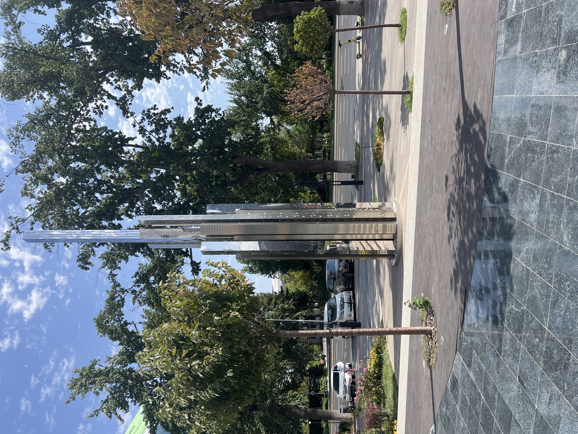 The image shows a modern sculpture in a city setting. The sculpture is tall and metallic, with a faceted design that reflects the surrounding environment. It stands on a paved area with trees and a street visible in the background. Cars are parked on the street, and a yellow electric scooter is on the sidewalk. The sky is blue with some clouds. Based on visual cues such as the architecture and vegetation, this scene is likely located in Tashkent, Uzbekistan, possibly along one of the city's main avenues or in a newly developed urban area.