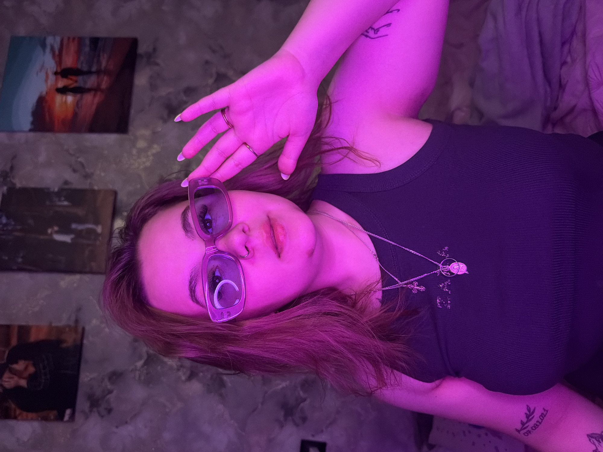 A young woman is positioned in front of a wall adorned with three paintings. She wears rectangular glasses, a dark tank top, and a silver necklace with circular pendants and a cross. She is lying down, with her right hand raised near her face. The room is illuminated with purple lighting. The location is not discernible from the image.