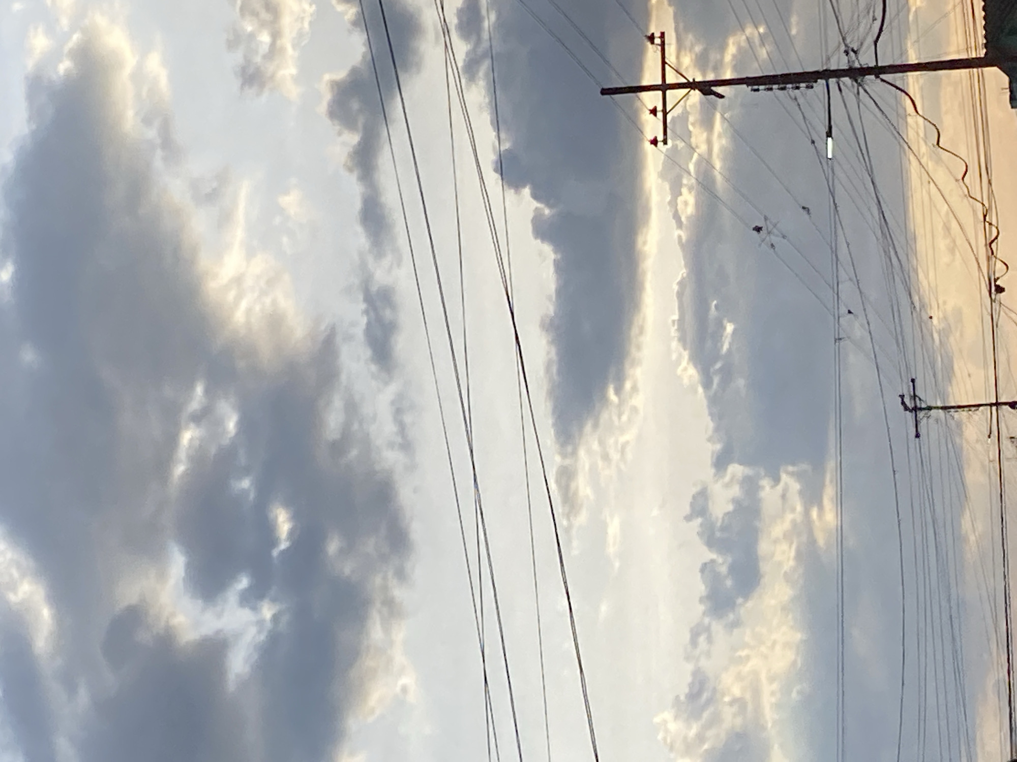 The image shows a sky with scattered gray and white clouds, partially illuminated by sunlight. Several utility poles with power lines crisscross the scene. The location is Tinaquillo, Venezuela, as indicated by the prompt. No people are visible. The primary subjects are the sky, clouds, and utility infrastructure. There are no evident actions or interactions occurring in the image. The environment appears to be an urban or suburban area, given the presence of utility poles. The image orientation is sideways.