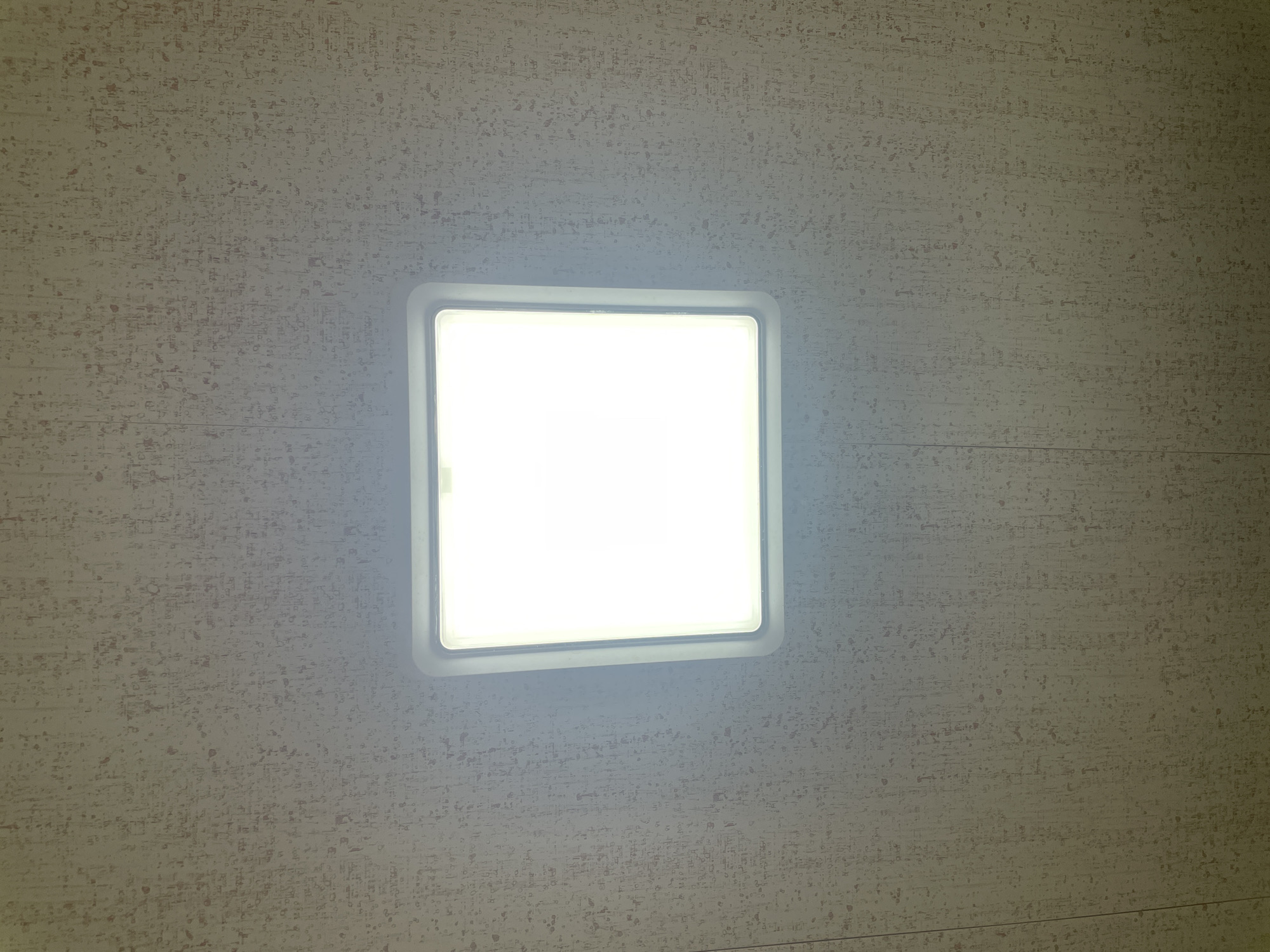 A square, white ceiling light is centered in the frame against a textured, neutral-colored ceiling. The ceiling appears to have a speckled pattern. The location is unspecified.