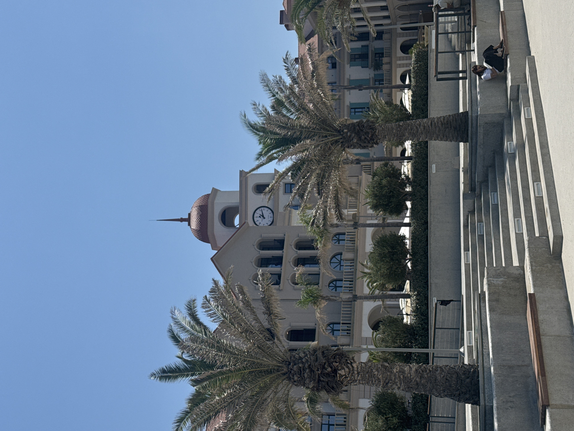 This image depicts an outdoor scene with a large, multi-story building as the backdrop. The building features a prominent clock tower with a red dome, multiple arched windows, and balconies. In the foreground, there are several palm trees, with two large ones framing the central part of the image. A tiered concrete structure with steps and benches is in the lower portion of the frame. Two individuals are visible on the steps; one is sitting and reading, and another person is partially visible in the background. The environment appears to be a public space, possibly a plaza or promenade, with a clear blue sky overhead. Based on architectural style and the presence of palm trees, this scene is likely located in a warm climate. Specifically, the building and its surroundings are identifiable as the Baku White City's Boulevard area in Sabunchu Raion, Azerbaijan.