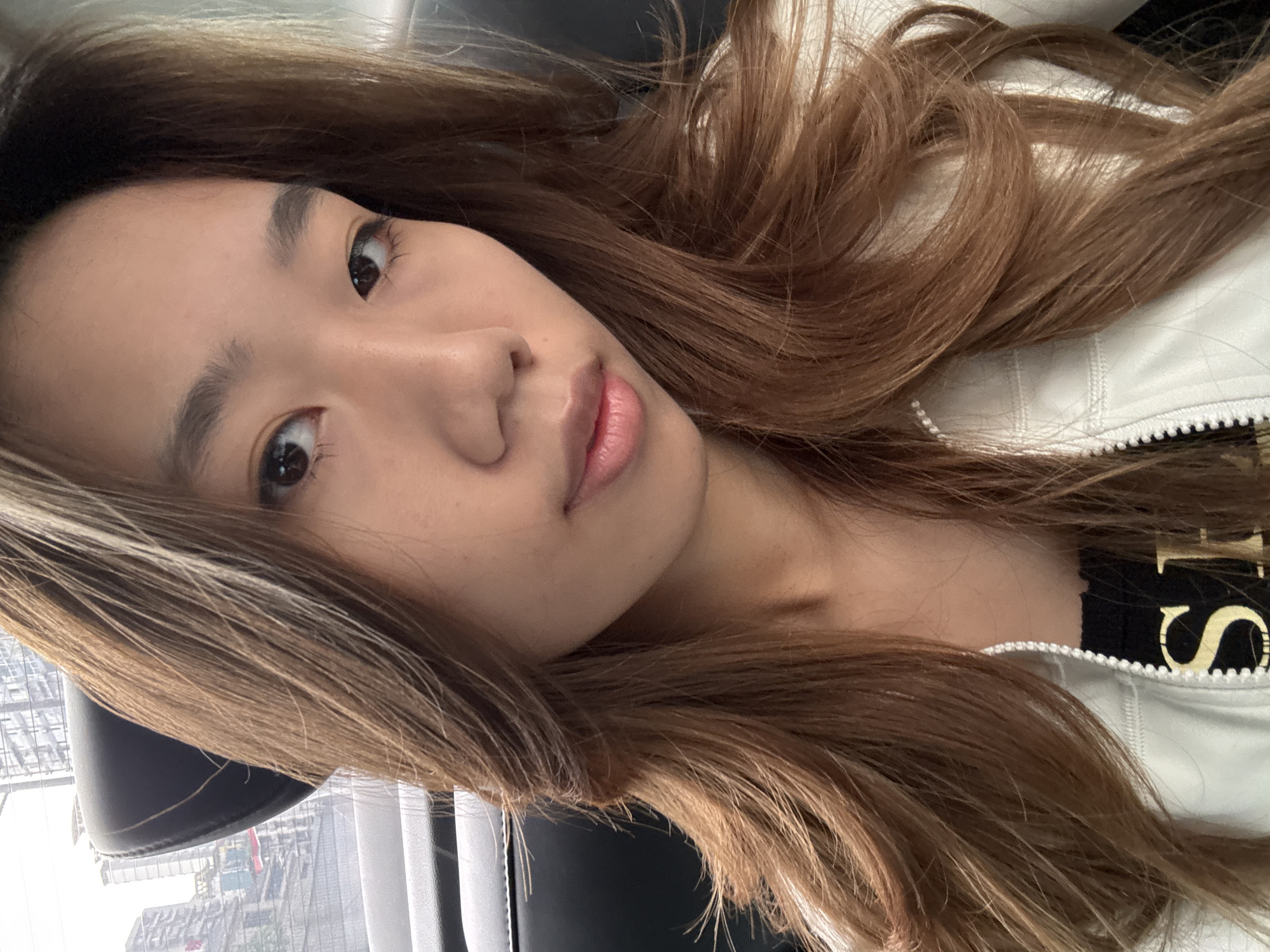 A close-up portrait of an Asian woman with light brown hair facing the camera at a slight angle. She is wearing a white jacket over a black shirt with gold lettering. The background appears to be a window with a view of a city skyline, possibly in Liangjiang, China. The scene is indoors, likely in a vehicle or building with a window.