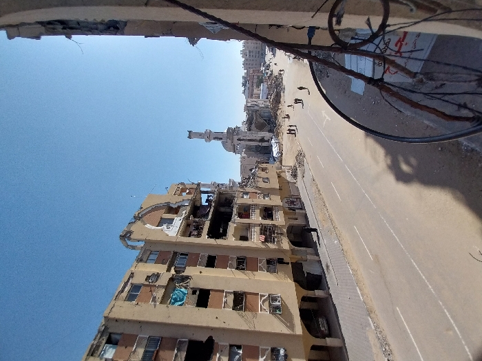 The image depicts a heavily damaged urban street scene in Khan Yunis, Palestinian Territory, under a clear blue sky. From an elevated perspective, a wide, paved two-lane road with white markings runs through the center. On the left, a multi-story, light-colored residential or commercial building stands severely damaged, with large sections of its facade missing, exposing interiors, broken walls, and empty window frames. Some balconies contain debris or fabric, and a light-colored sedan is parked beneath an arched ground-floor section. A single individual in a black abaya walks on the sidewalk adjacent to this building.
In the mid-ground, a group of approximately 5-7 people, mostly in dark clothing, walk along the street and amidst rubble on the right side. Beyond the street on the right, substantial piles of debris indicate demolished structures. A prominent, light-colored mosque with a tall, slender minaret is visible in the background, showing damage to its dome and upper sections. The foreground on the right is framed by a partial view of a damaged concrete structure, possibly a balcony, with exposed rebar and a metal railing. A partially obscured white sign with Arabic script is visible behind the railing. In the distance, other urban buildings, some appearing damaged, complete the skyline.