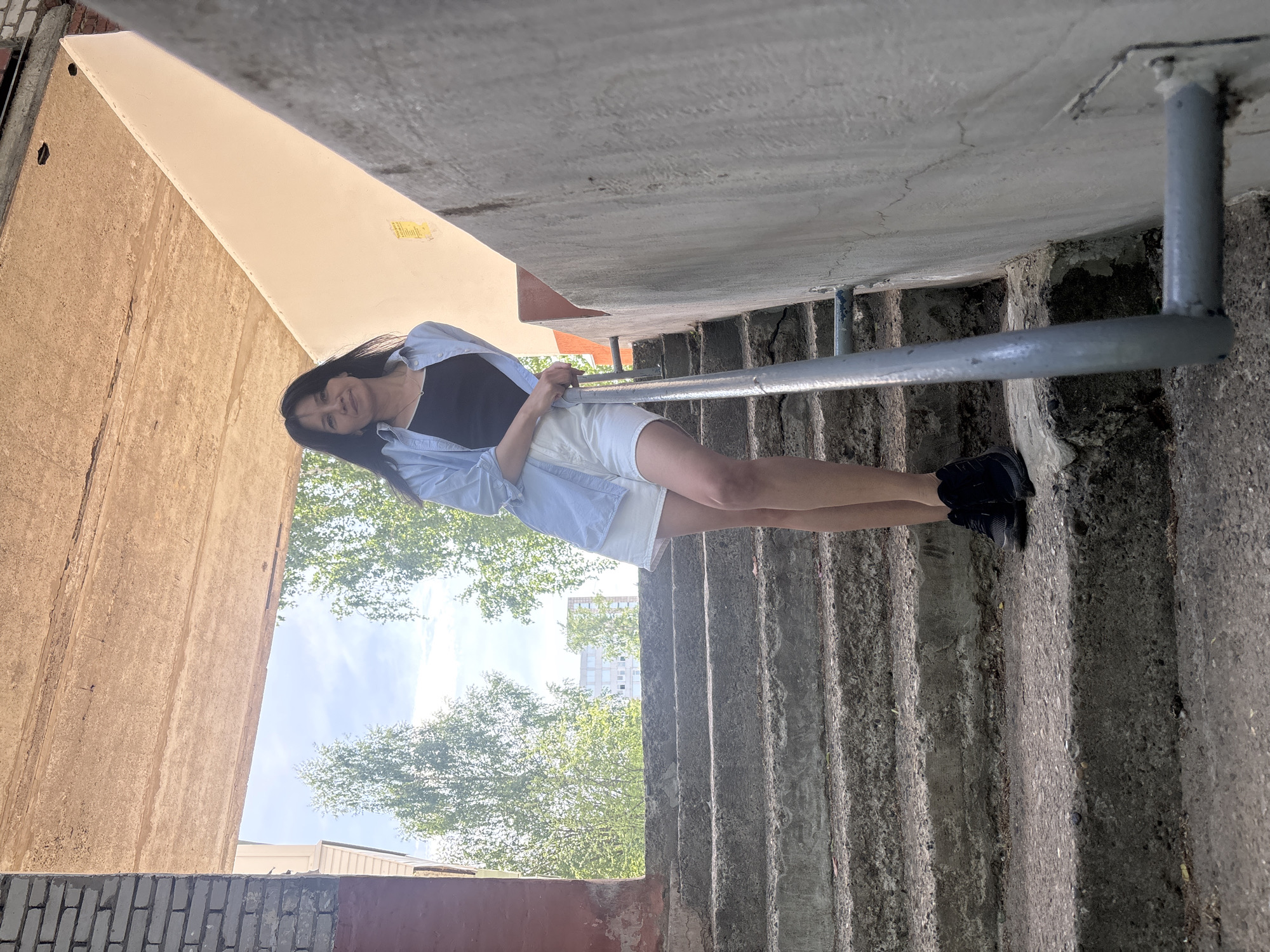 The image shows a woman posing on a staircase in what appears to be an urban environment. She is standing on concrete steps with a metal railing, wearing a black top, a light blue open shirt, and white shorts. She is leaning against the railing and looking towards the camera.
In the background, there are trees and a glimpse of the sky. There is a concrete structure above the staircase and a brick wall on the left side of the frame.