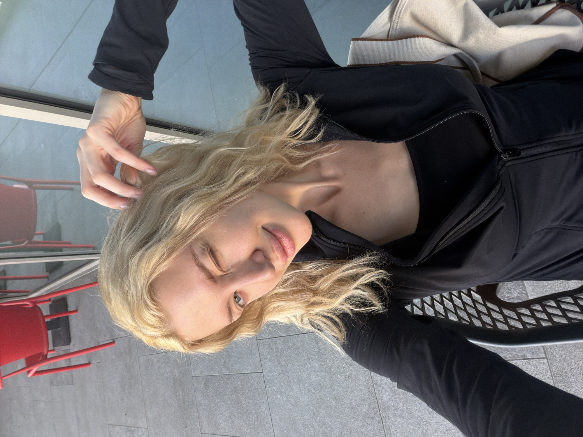 A young woman with blonde hair is lying on a metal bench in what appears to be an outdoor public space. She is wearing a black zip-up jacket over a black top. Her left hand is near her hair, and she is looking at the camera with a slight smile. To the left of the bench, red chairs are visible in a window reflection. The ground is paved with light grey tiles. A light-colored bag is partially visible next to her upper body. There is no definitive information to confirm that the image was taken in Belgrade, Serbia.