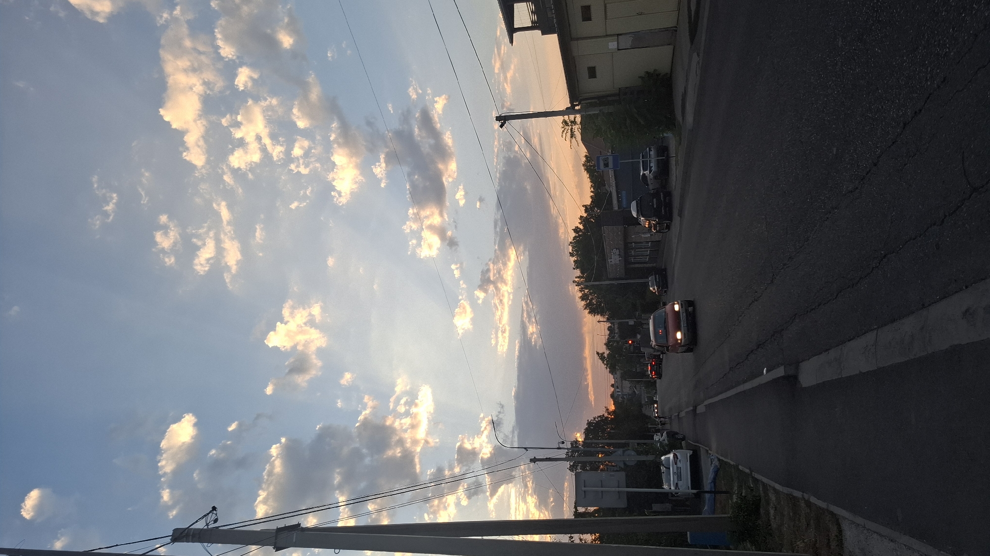 The image shows a street scene in Shymkent, Kazakhstan, during what appears to be either sunrise or sunset. The sky is filled with scattered clouds, some illuminated by warm, golden light, suggesting either dawn or dusk. The foreground features a paved road with visible cracks and texture, flanked by a sidewalk on the left. Several cars are present on the road, with some in motion and others parked.
On the left side of the road, a tall utility pole with wires is prominent, along with a few trees and a building. The sidewalk has some debris and a blue cylindrical object on its edge. Further down the street, more buildings are visible, including what looks like a shop or business with signage. On the right side of the road, another utility pole and a low-rise building with a yellow facade are present. The overall environment appears to be a residential or mixed-use urban area. There are no clearly identifiable people in the image, though some indistinct figures might be present near the parked white car on the left. The lighting conditions indicate a natural light environment, likely outdoors.