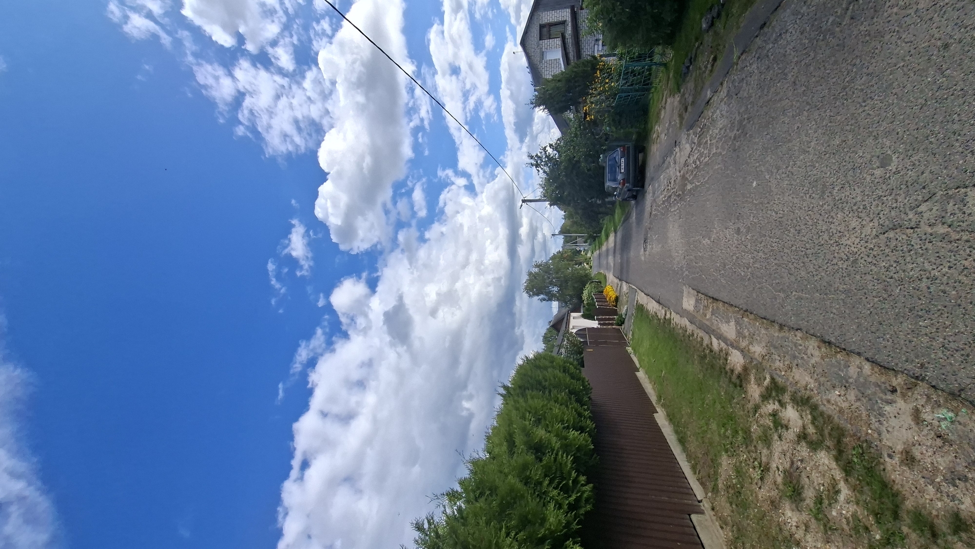 The image depicts a sunlit rural road in Myadzyel, Belarus, under a bright blue sky dotted with fluffy white clouds. A dark brown wooden fence lines the left side of the road, topped by a dense row of green evergreen trees. Beyond the fence, a house with a white facade is visible. The asphalt road is in moderate condition, with some patching and unevenness. A dark-colored car is parked on the right side of the road. Further down the road, houses and lush green trees line both sides, with some yellow flowers visible in a garden. The scene suggests a peaceful summer day in a suburban or village setting.