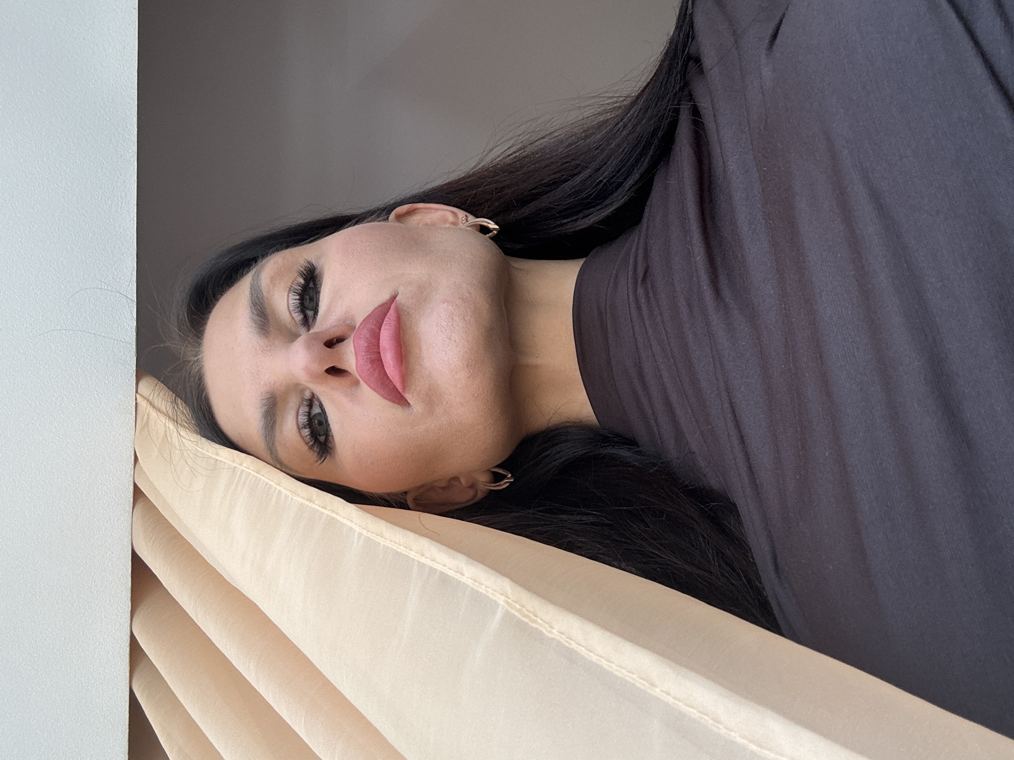 A woman with dark hair is lying down next to a wall and a peach-colored curtain. She is wearing earrings and a dark-colored top. The picture appears to have been taken at an angle looking down on the woman. The location information is not discernible from the image alone.