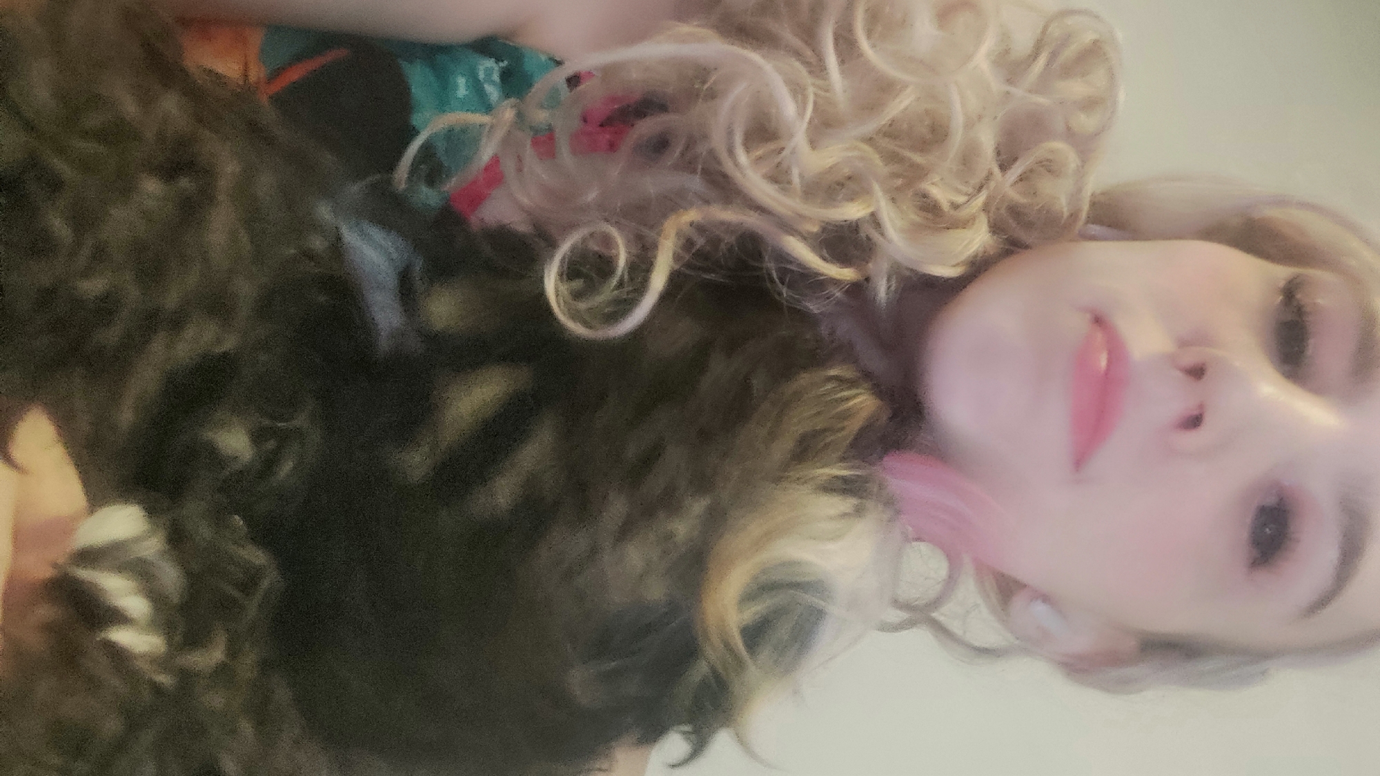 A selfie shows a fair-skinned individual with curly, blonde hair, fair features, and pink lips, positioned against a light-colored background. Beside the person is a black, long-haired animal, possibly a dog, with a white-tipped paw visible. The image is taken at a close range, and the location is unknown and cannot be ascertained from the image.