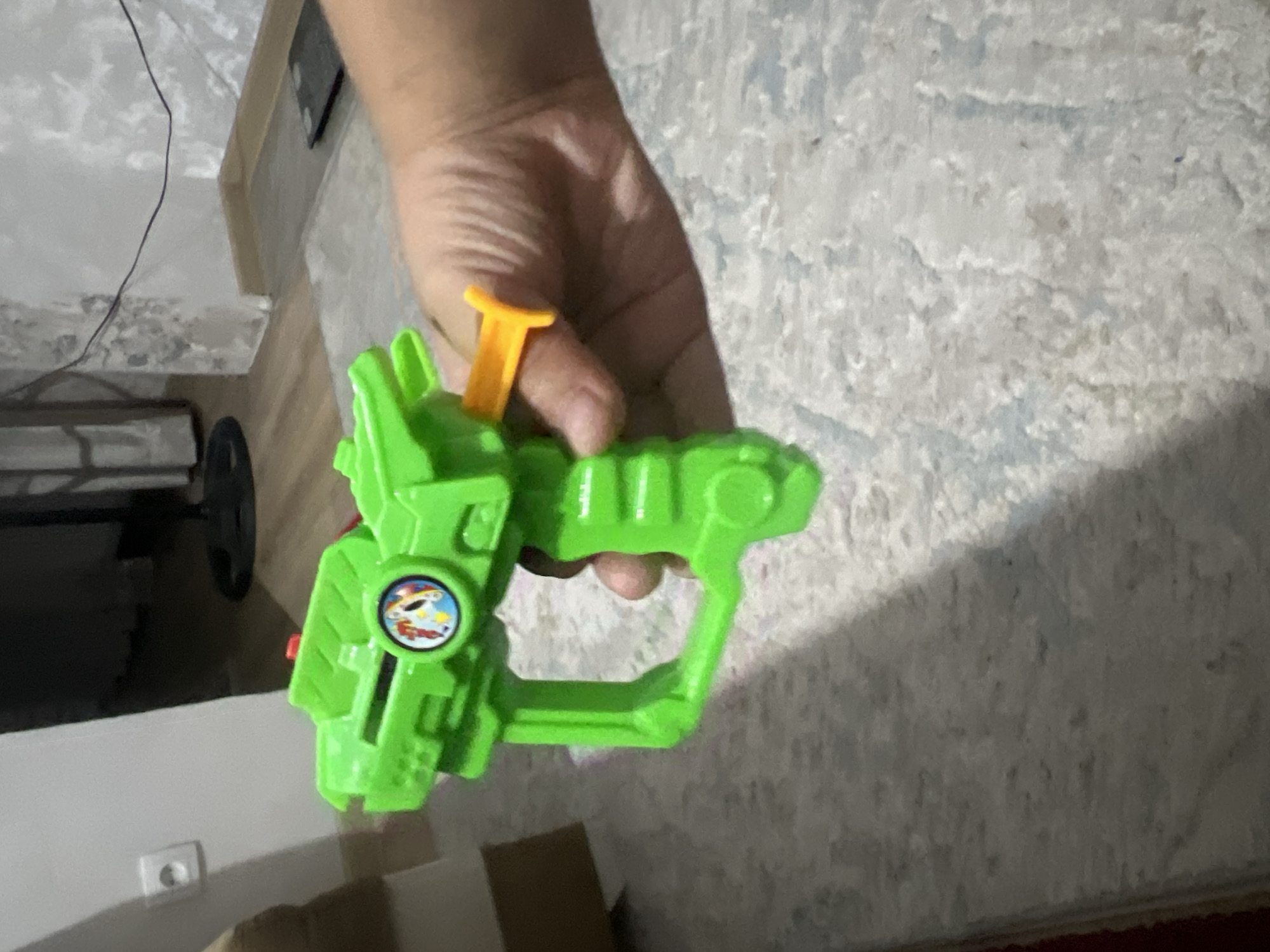 An overhead, medium shot captures a bright green plastic toy gun held by a person's hand. The toy gun, with a yellow trigger, is the primary focus, positioned in the lower center of the frame. The background consists of a light-colored carpeted floor and a textured wall with faint patterns. A white electrical outlet is visible on the left side of the wall, near a blurred brown object that could be furniture. The lighting appears to be natural, coming from an unseen source, casting soft shadows. The overall scene is indoors, in an environment that suggests a domestic setting.