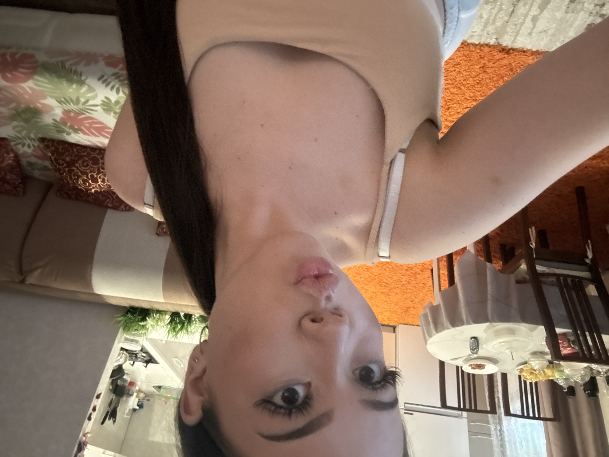 The image displays a selfie taken by a young woman in an indoor home setting, likely in Naberezhnye Chelny, Russia. The woman, positioned in the foreground, is looking upwards towards the camera with a playful expression, featuring pursed lips and wide eyes accentuated by prominent false eyelashes and dark eyeliner. Her long, dark hair cascades over her left shoulder. She is wearing a light beige or nude-colored tank top with thin white bra straps visible. Several small moles are scattered across her upper chest.
The background reveals a domestic interior. To the left, a brown sofa is partially visible, adorned with patterned cushions, including one with a red and gold geometric design and another with a pink and green floral motif. To the right, a wooden dining table is set with chairs, plates, a remote control, and some food items, such as golden-brown snacks and a dish containing reddish contents. The floor is covered by an orange and cream geometric patterned rug. Artificial green plants add a touch of decor, and a reflection of a kitchen area with a sink and taps can be faintly seen on a glossy surface. The lighting is bright and even, indicating a well-lit indoor space, likely during the daytime.