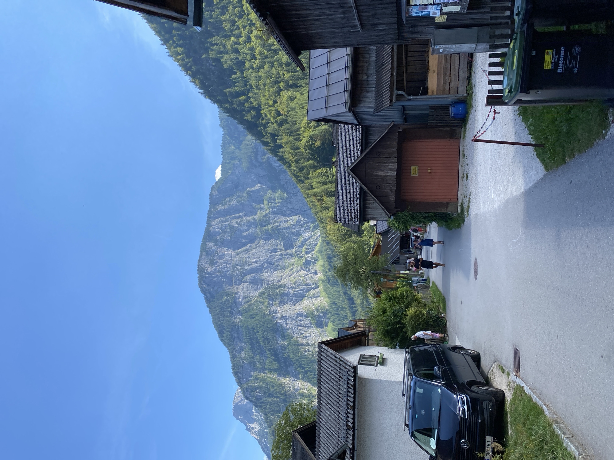 The image depicts a street scene in Hallstatt, Austria, with a focus on traditional architecture and the imposing mountain backdrop. The foreground shows a black Volkswagen van parked on the left side of a paved road, with grass and a curb bordering the vehicle. A man in a white shirt and pink shorts is standing near the van. Further down the road, two people are walking away from the camera. On the right side of the road, a series of wooden buildings with sloped roofs are visible, some appearing to be garages or storage sheds. One building has a prominent red door. The background is dominated by a large, steep mountain covered in lush green trees, with rocky cliffs visible towards the summit. The sky is clear and blue. There are also two green trash bins with yellow labels on the right side of the frame, near a fence.