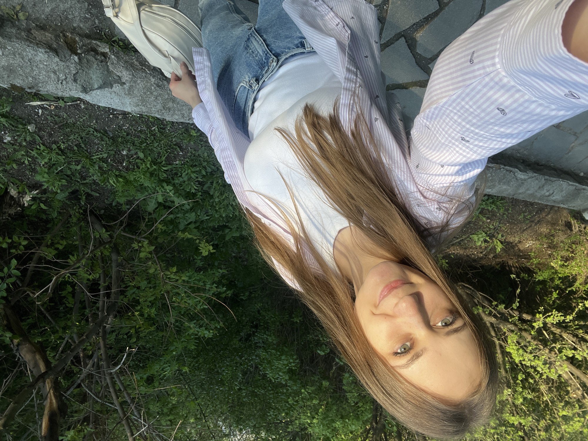 A young woman is centered in the foreground, taking a selfie. She has long, light brown hair, fair skin, and green eyes. She is wearing a white tank top, light blue jeans, and a pink and white striped shirt over the tank top. She holds a cream-colored purse. The background includes greenery and foliage of bushes and trees, with a cobbled stone path and raised curb. The photo gives the impression of an outdoor setting. The location cannot be identified as Yekaterinburg, Russia, without additional context or visual cues specific to that city.
