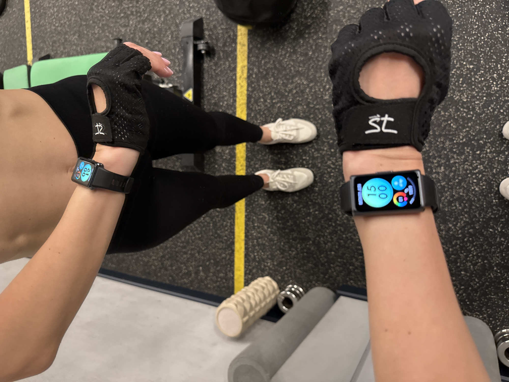 The image shows a first-person perspective of two individuals in what appears to be a gym. Both individuals are wearing black fitness gloves and black smartwatches. They are dressed in black leggings and white sneakers. The gym floor has a dark, speckled texture, with a yellow stripe. Exercise equipment, including foam rollers and weights, are visible. There is no information to determine that the location is in Sochi, Russia.