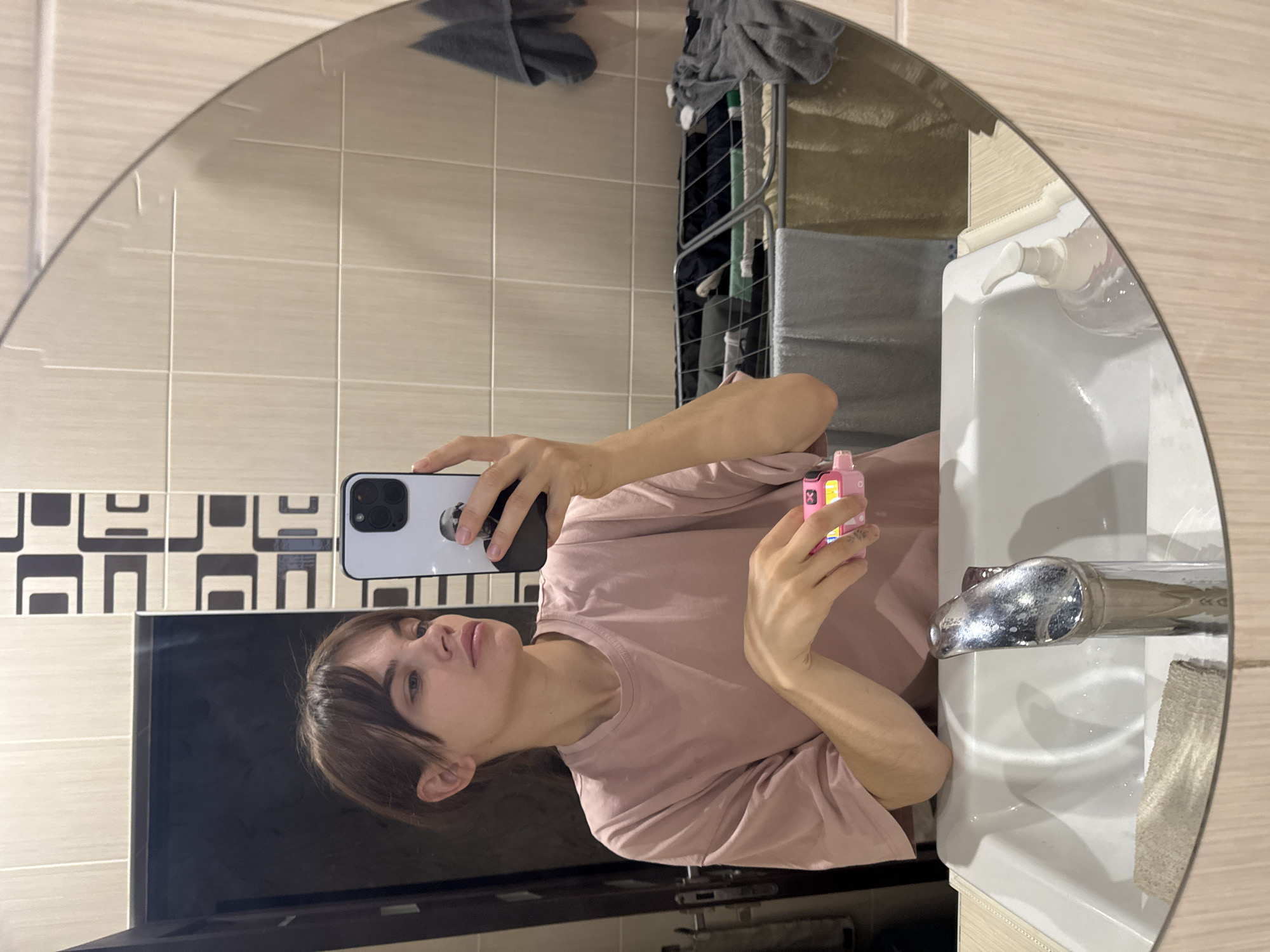 This image is a mirror selfie taken by a person in a bathroom. The subject, who appears to be female with light brown hair, is captured from the chest up, wearing a light pink t-shirt. She holds a white smartphone, likely an iPhone due to its camera design, with her left hand to take the photo. The phone has a dark case and a white circular pop socket or stand on its back. In her right hand, she holds a small, rectangular pink device, which is identifiable by the prominent white text "VOOPOO" and "ARGUS G" as a VOOPOO Argus G vape.
The bathroom features light beige tiled walls, with a section of decorative black and white geometric pattern tiles on the left. A white ceramic sink with a shining chrome faucet and a white pump-style soap dispenser are visible on the right. In the upper background, a metallic clothes drying rack with grey towels or clothing hanging on it can be seen. Below the patterned tiles on the left, a large, dark, reflective surface, possibly a television or monitor, is partially visible.
The lighting suggests an indoor setting, likely with artificial light, as no natural light sources are apparent. The person's expression is neutral. There is no weather information visible. The location context provided is Rostov-on-Don, Russia.