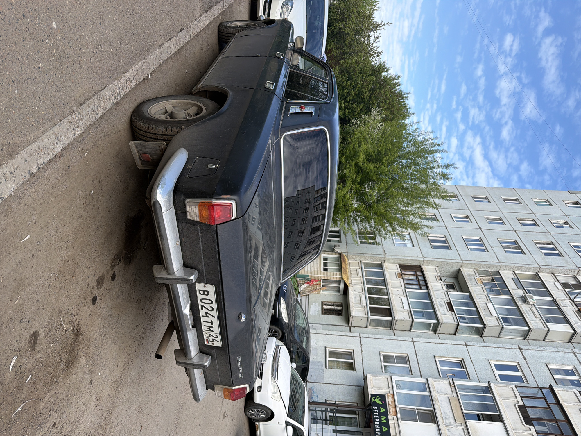 A black GAZ-24 Volga is parked on a street in Krasnoyarsk, Russia. The license plate reads "B 024 TM 24" and indicates that it is a vehicle licensed in the Krasnoyarsk region. The car is located on the side of the road next to a sidewalk and in front of a large, gray, multi-story building. There are trees between the building and the car. A white car is parked ahead of the Volga.