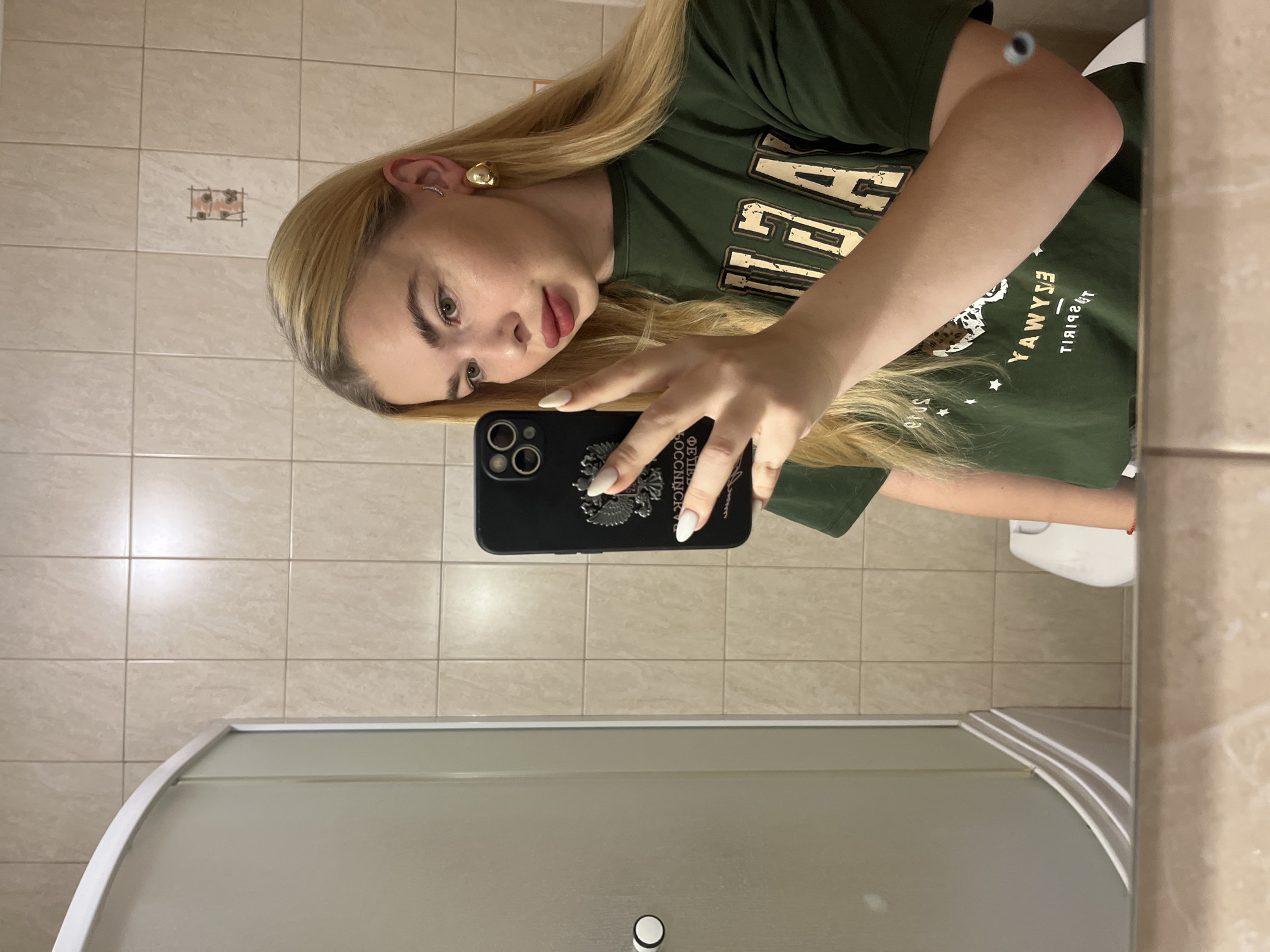 A young Caucasian woman with blonde hair is taking a mirror selfie in a bathroom. She is wearing a dark green t-shirt with text and graphics on it, and large gold hoop earrings. She has on a light-colored lipstick and her nails are painted white. The bathroom has tiled walls and a glass shower enclosure. A toilet is partially visible in the reflection of the mirror. The scene is not distinguishable as being in Krasnodar, Russia.
