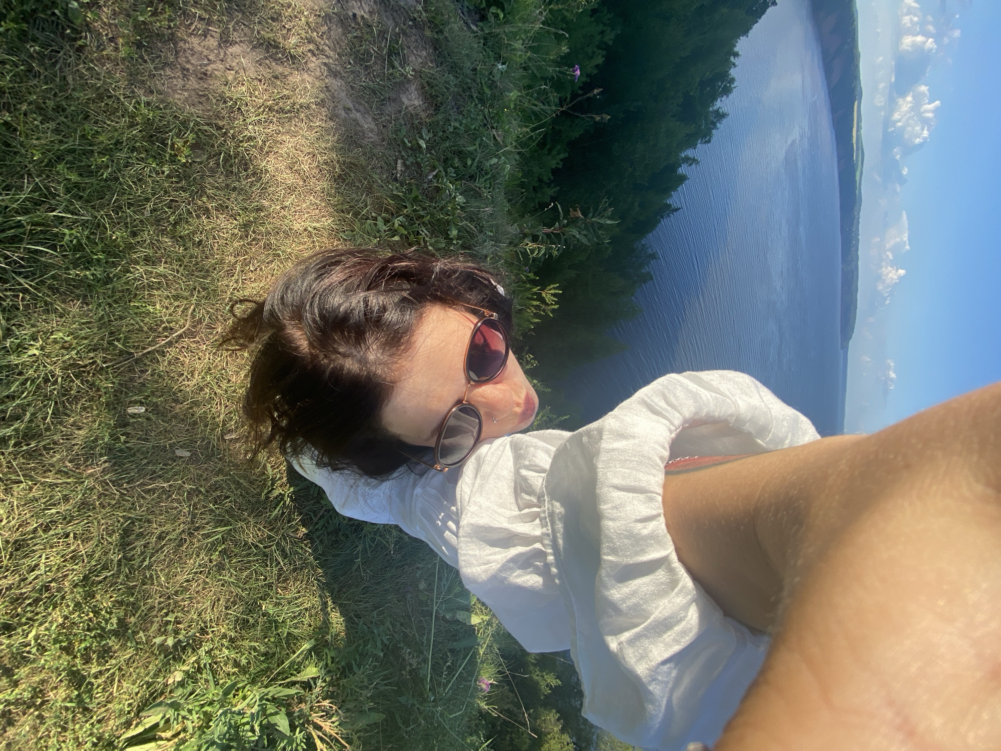The image shows a woman with dark hair, wearing sunglasses and a white dress, positioned upside down on a grassy cliff overlooking a body of water. The location is the city of Kostovaty, Russia. The woman's arm is visible in the foreground, obscuring part of the frame. The background features a vast expanse of blue water, with green, tree-covered hills and a clear sky with clouds. The terrain is covered in green grass and vegetation.