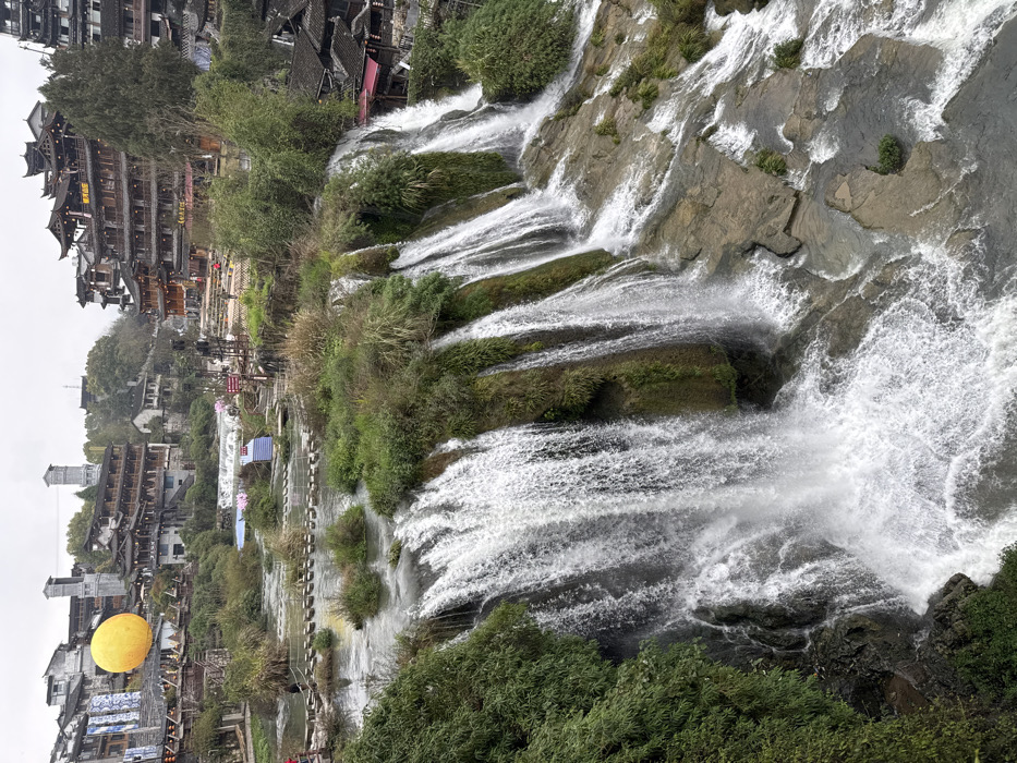 The image captures a panoramic view of the Furong waterfall and surrounding ancient town in Furong, Hunan, China, under an overcast sky. A wide waterfall dominates the foreground and mid-ground, with numerous cascades of white water flowing over a multi-tiered, moss-covered rock formation into a turbulent pool below. Above the waterfall, a river flows, traversed by a line of approximately twenty irregular, dark grey stepping stones. Several individuals are visible on or near these stones, with one person in a red top standing on the left bank.
The riverbanks, small islands, and parts of the cliff face are covered in lush green and brown vegetation, including shrubs and grasses. Traditional multi-story Chinese buildings, characterized by dark tiled roofs and wooden structures, are built into the hillsides in the mid-ground and background on both sides of the river. On the left side, a large, bright yellow spherical object is positioned above the rooftops, with two grey, square-sectioned towers visible further in the background. On the right side, additional traditional buildings with curved eaves and red architectural accents are clustered. Two small blue tarpaulins are visible on the right bank above the waterfall. Pedestrian pathways are discernible winding through the town structures.