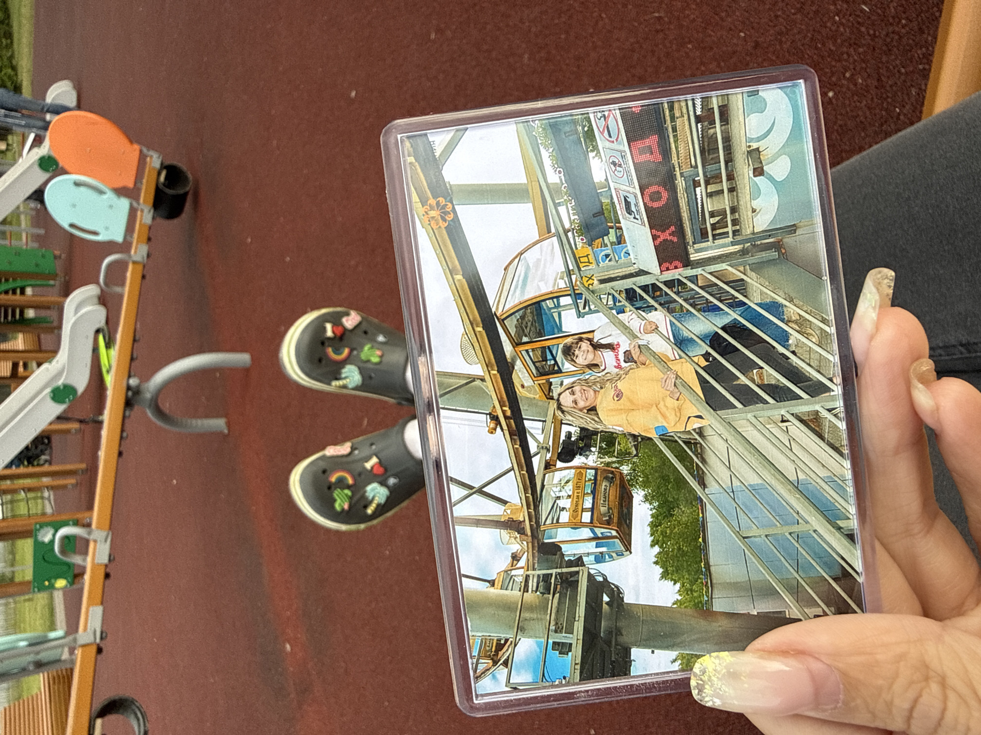 A hand, adorned with light-colored, glittery nail polish, holds a clear plastic card displaying a photograph. The photograph shows two women riding in a gondola of a Ferris wheel. One woman, with long blonde hair, wears a yellow hoodie, while the other, with darker hair, wears a white top with red lettering, possibly "BOUY". Both are smiling and appear to be posing.
The Ferris wheel in the photo has classic-style gondolas, one of which displays "Ferris wheel 1911" and "Иваново" (Ivanovo), suggesting the location and possibly age of the attraction. Other details within the photo include parts of the Ferris wheel structure, other gondolas, trees, and a building with prominent red "BOX" lettering and other signs containing symbols. The sky appears bright, indicating daytime.
The hand holding the card is positioned over a reddish-brown, textured ground, likely a rubberized playground surface. Two feet wearing black Crocs-style shoes, adorned with various charms including a rainbow, a heart, a palm tree, and "I <3", are visible on the ground. Partially visible in the background is playground equipment, including light blue and orange seats on a stand, a silver slide, and other green and metallic structures. A segment of a leg in dark pants is also seen. The overall scene suggests an outdoor setting, specifically a playground in Шебунино, Russia, where the photo of the hand holding the card was taken.
