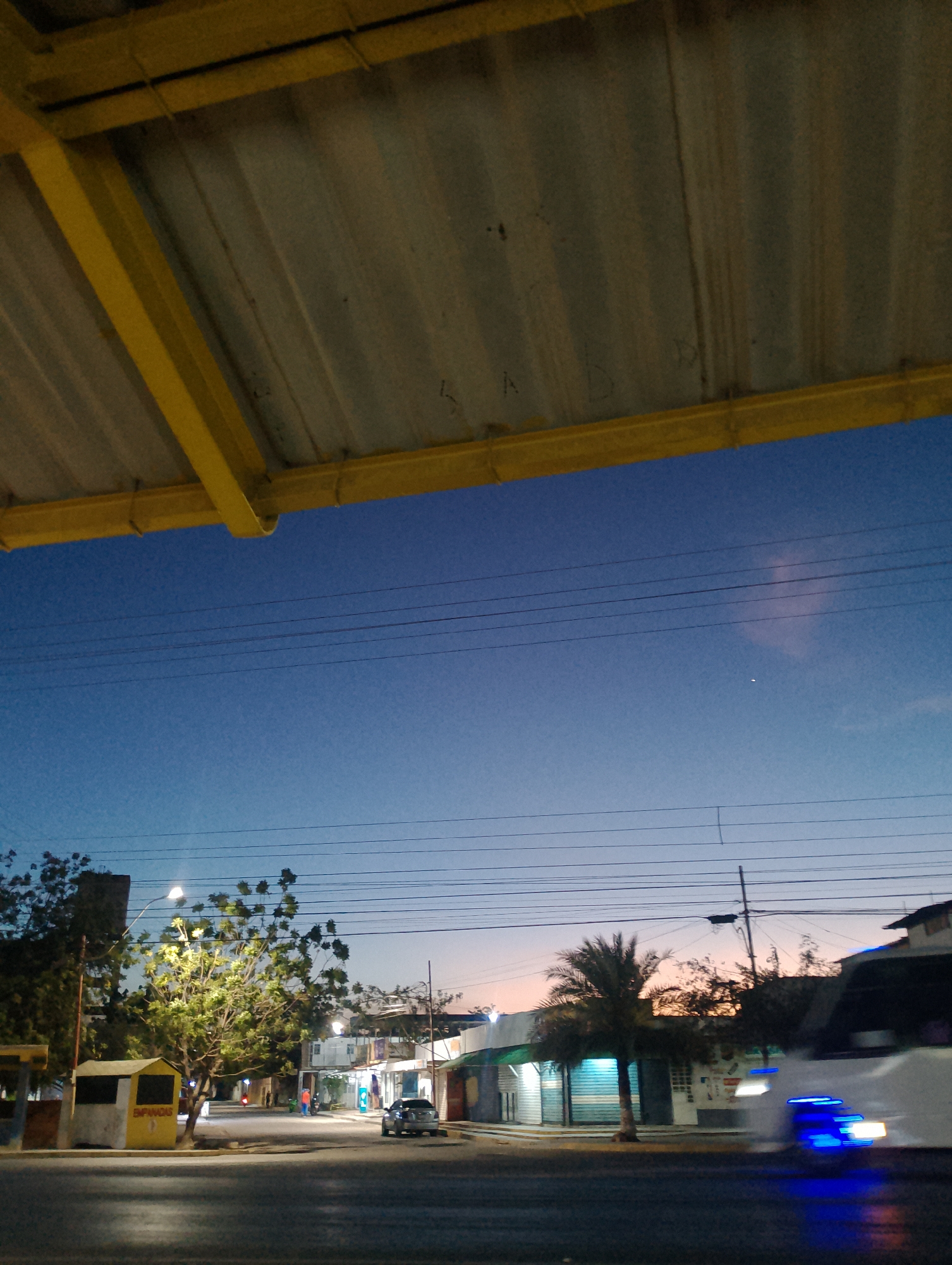 The image is a low-angle shot taken at dusk or dawn in Las Barrancas, Venezuela. An overhead structure, likely a bus stop or awning with a yellow frame and corrugated metal roofing, dominates the upper portion of the frame, with some graffiti etched on the roofing.
Below, the sky transitions from a deep blue at the top to a light orange glow near the horizon. Several power lines run horizontally across the sky.
On the ground, the scene depicts a street. A small yellow "EMPANADAS" stand sits to the left. Trees with bright street lighting surround it.
Across the street, storefronts are visible, with a palm tree punctuating the scene. A vehicle with its headlights illuminated blue is blurred by motion in the foreground of the image, moving to the right. A second vehicle is visible parked on the left side of the street. There are at least two people visible in orange clothing in the middle of the cross street.