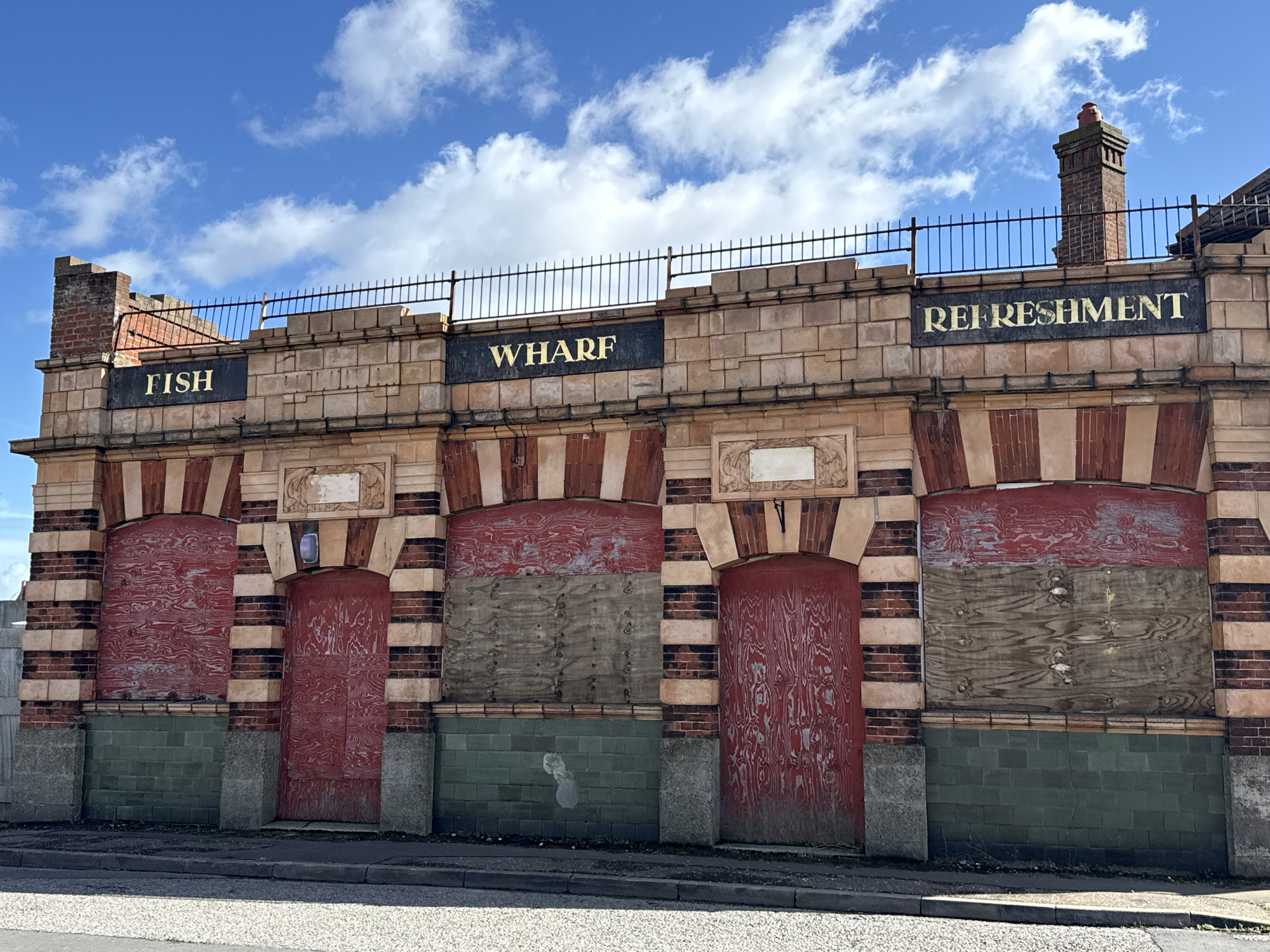 A disused, single-story brick and stone facade of a former commercial building is depicted under a partly cloudy blue sky in Gorleston-on-Sea, United Kingdom. The structure features a horizontal upper frieze with three black rectangular signs bearing gold-colored sans-serif text: "FISH" on the left, "WHARF" in the center, and "REFRESHMENT" on the right. Above the frieze, a dark metal railing with vertical balusters runs across the top, punctuated by two brick chimneys on the left and right ends, the rightmost chimney topped with a terracotta pot.
The building's lower section presents four arched openings, alternating between wider windows and narrower doorways. These openings are framed by alternating courses of light beige stone and red brick. Three of the openings are boarded up with plywood sheets. The leftmost and rightmost arched openings are covered with plywood painted a faded, peeling red, revealing the wood grain beneath. The two central arched windows are covered with unpainted, weathered plywood sheets, showing visible nail holes and horizontal seams. Decorative stone panels with faded bas-relief details are positioned above the arches between the openings.
The lower half of the facade, beneath the arched openings, is faced with dark green glazed tiles, showing some discoloration and damage. Concrete plinths support the tile work beneath the brick and stone columns separating the arches. The building sits directly adjacent to a paved public sidewalk and a dark asphalt street, visible in the foreground. No people or active interactions are present in the scene. The overall condition suggests a state of disrepair and abandonment.