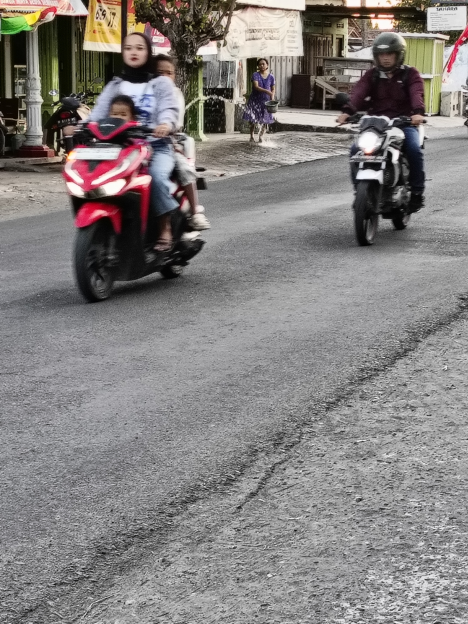 In Sukorini, Indonesia, a woman wearing a hijab and a grey jacket is riding a red scooter. She is accompanied by two children, one in front and one behind her. The woman is looking forward, and the children are also looking forward. The scooter is moving forward on a paved road. In the background, there are buildings and trees. To the right, a man in a maroon jacket and helmet is riding a white motorcycle. A woman in a purple dress is standing on the left side of the road holding a bucket.