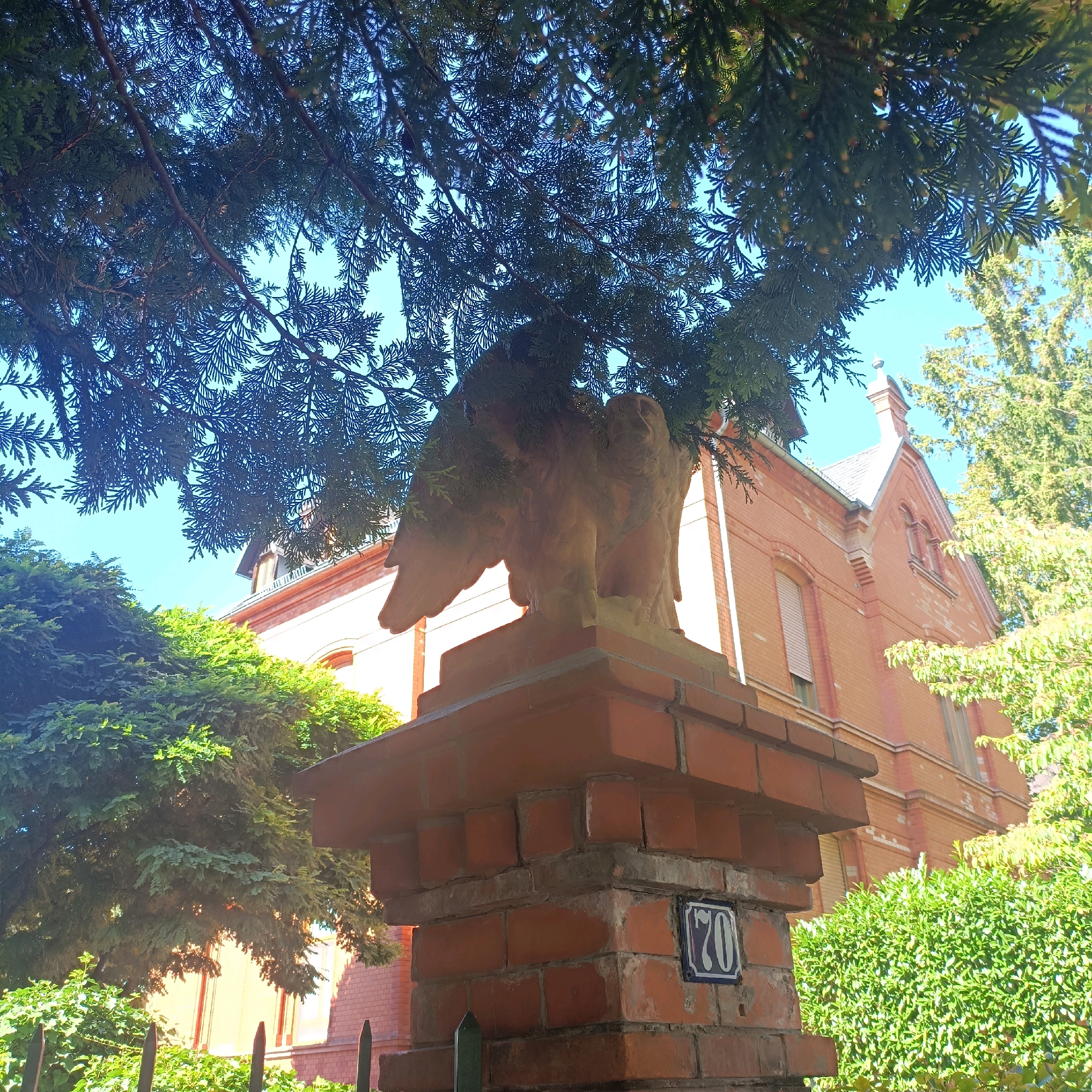 The image shows a terracotta eagle sculpture perched atop a brick pillar in front of a red brick building. The pillar has a blue and white house number "70" sign on it. Lush green trees and foliage surround the pillar and building. The scene appears to be a residential area. The style of the building and the house number plate suggest it is located in Germany, possibly in a city like Wiesbaden, which has many historic buildings with similar architectural features. The image captures a bright, sunny day with clear blue skies visible through the trees.