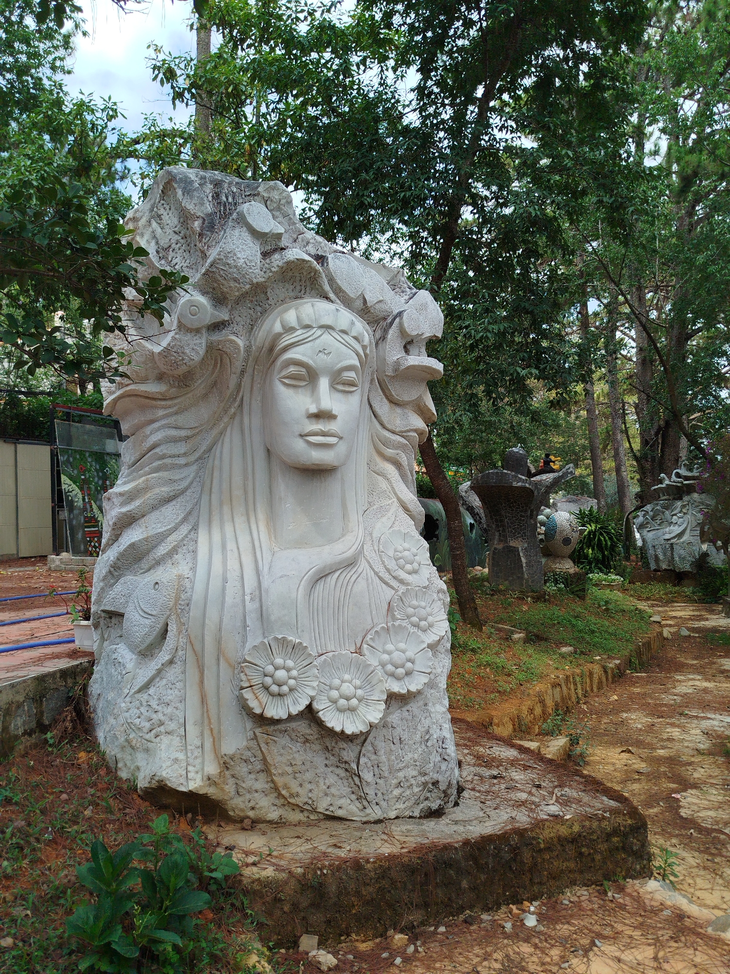 The image shows a large, white stone sculpture of a woman's bust in a garden setting in Da Lat, Vietnam. The sculpture features floral details around the neck and flowing hair-like carvings. Other sculptures are visible in the background, along a winding path covered in fallen leaves and red dirt. Lush green trees surround the statues, creating a partially shaded environment. The sculpture is positioned on a mossy, stone base. A building is partially visible in the background to the left.