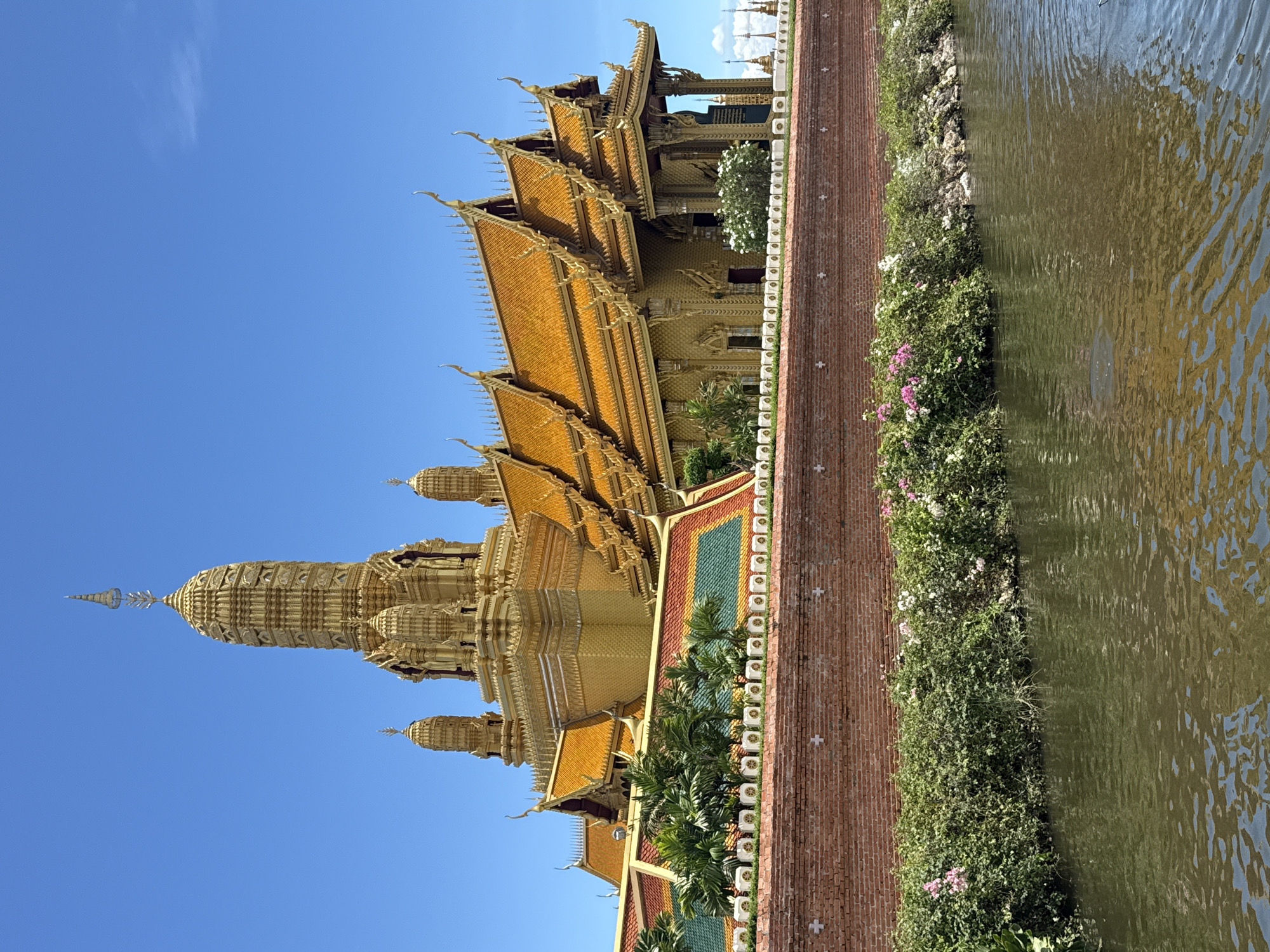 A golden temple complex sits beside a waterway in Bang Pu Subdistrict Municipality, Thailand. The temple boasts multiple tiers of ornate golden roofs, with a central spire reaching toward a clear blue sky. A brick walkway runs along the edge of the temple grounds, separated from the waterway by a strip of vegetation, including flowering plants. The water surface reflects sunlight.