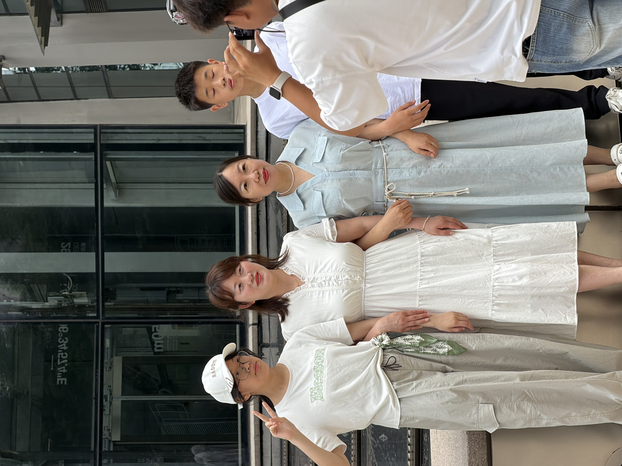 In Yuzhong District, China, four individuals are posing for a photograph in front of a modern building with large glass windows reflecting the sky and trees. On the left, a young woman wearing a white cap, glasses, and a white t-shirt with green lettering that reads "Blesson," and light-colored cargo pants, is making a peace sign with her left hand. Next to her stands a woman in a white, ruffled-neck dress. To her right, a woman in a light blue dress with a pearl necklace and a braided belt is also posing. Behind them, a young man in a white t-shirt and black pants stands with his eyes closed. The photographer, a person wearing a white t-shirt and jeans, is partially visible in the foreground, holding a camera to take the picture. The individuals are standing close together, with their arms around each other, suggesting a family or group outing.