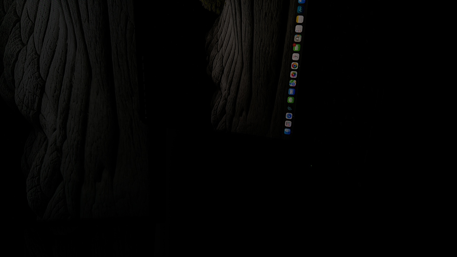 The image shows a close-up of a dark, abstract landscape, possibly rock formations or rippling water, with a portion of a computer screen visible in the lower right. The screen displays a dark wallpaper with a dock of application icons at the bottom. The icons are small and brightly colored, including what appears to be a Safari browser icon, a mail icon, and a calendar icon. The overall scene suggests a dark, indoor environment with a focus on digital display. There is no visible connection to Kafr Yasif, Israel.