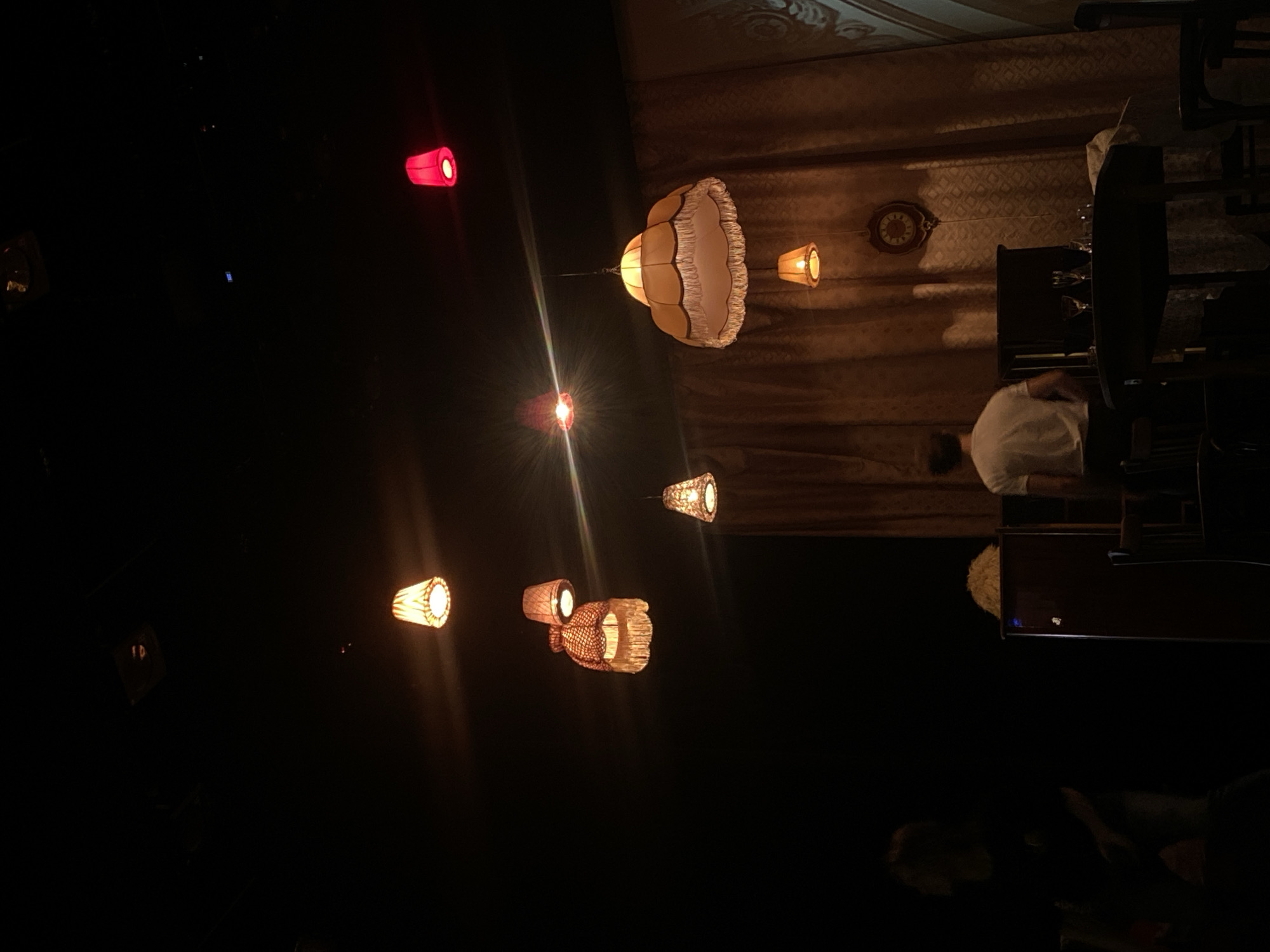 The image depicts the interior of a dimly lit space, likely a bar or lounge, in Zelenograd, Russia. Several decorative lamps of various shapes and sizes are suspended from the ceiling, providing the primary source of illumination. These lamps cast a warm, yellow glow throughout the room. A person wearing a white shirt is seated at a piano, seemingly playing. Behind the piano, a patterned curtain or wall covering hangs, and a clock is visible on the wall. The overall ambiance is subdued and atmospheric. A red lamp is visible in the distance. The bottom of the photo shows a partially visible person.