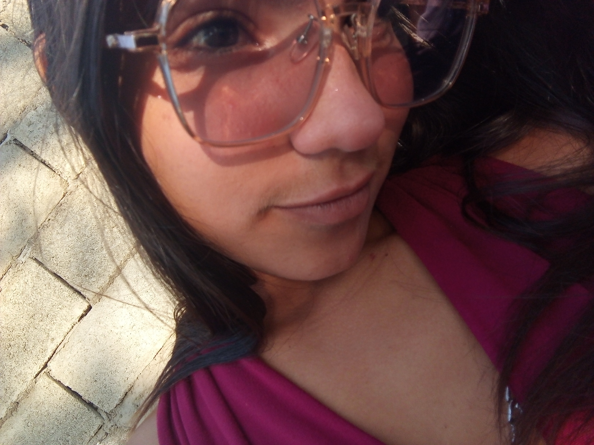 A close-up, slightly tilted selfie shot of a person with long dark hair and fair skin. The person is wearing large, transparent glasses with a metal frame, which cast shadows on their face. They are wearing a magenta top with a gray trim. The background is a blurred surface of gray paving stones, indicating an outdoor location. There is no explicit evidence or context to indicate the specific location as Juan Griego, Venezuela.