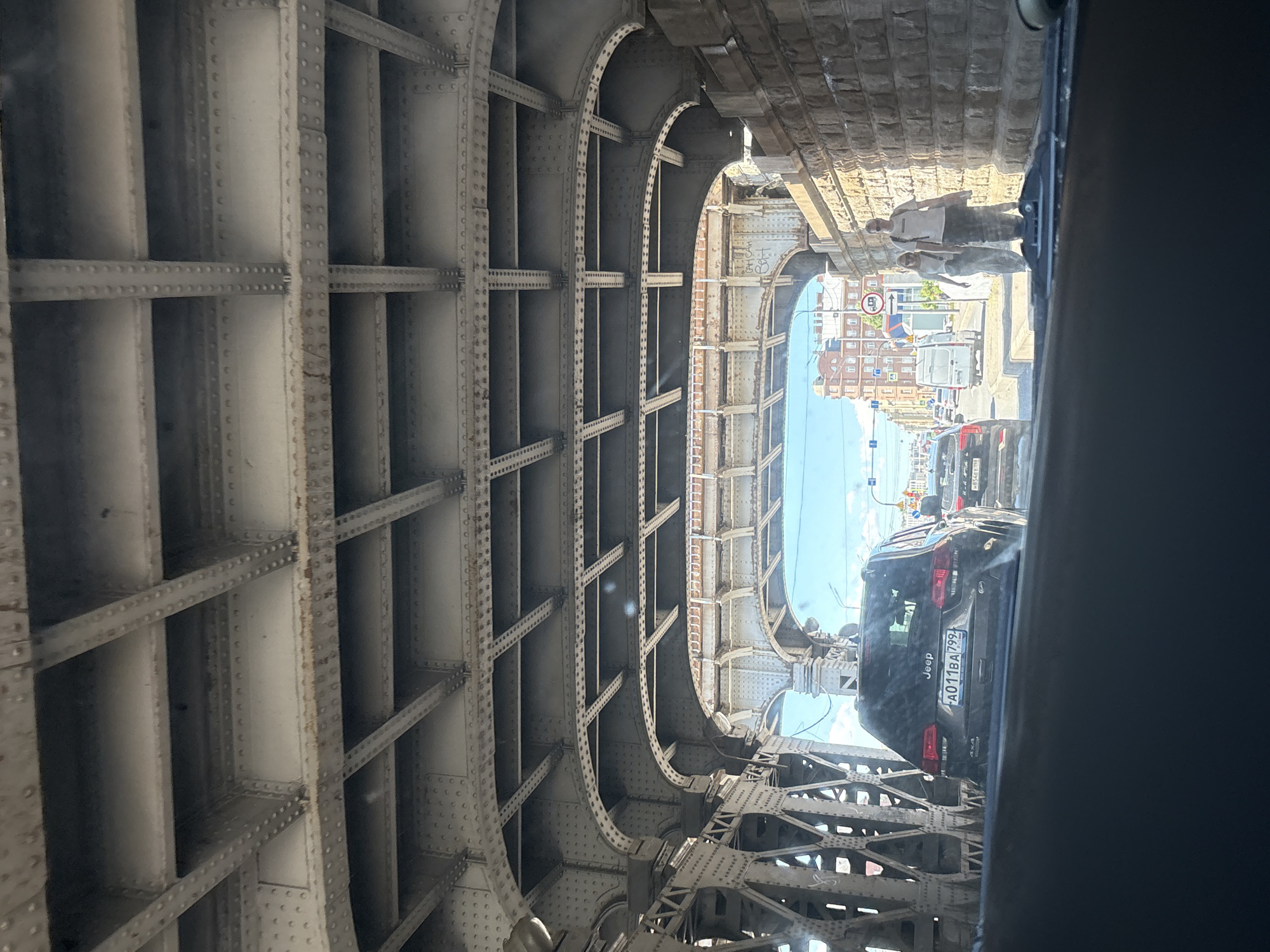 This is a photograph taken from inside a vehicle driving under a large, riveted metal bridge in Saint Petersburg, Russia. The view looks forward, through the arch of the bridge and out into a street scene. The foreground is dominated by the dark interior of the vehicle, including part of the dashboard and windshield.
The bridge structure itself is a prominent element, with intricate metalwork, girders, and crossbeams visible above. The color of the metal appears to be a weathered grey. On the right side of the frame, a stone wall is visible, forming part of the bridge's support structure.
Beyond the bridge, the street is visible. There are several cars on the road, including a dark-colored SUV with the Jeep logo prominently displayed on the back. A white van is also visible further down the street. Two people, a man and a woman, are walking on the sidewalk to the right of the road, near the stone wall. The man is taller and wearing a white t-shirt and dark pants, and the woman is wearing a light-colored dress and sunglasses.
In the distance, buildings with red brick facades and arched windows are visible, characteristic of Saint Petersburg architecture. The sky is partly cloudy and blue. Traffic lights and street signs can be seen on the left side of the street. The overall impression is a sunny day in an urban environment. The license plate of the Jeep is visible as A011BA799.