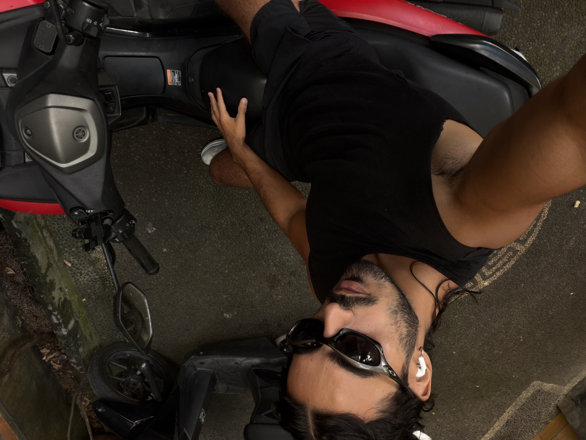 A man is photographed in an overhead, selfie-style shot, sitting astride a black and red Yamaha scooter. The scooter is parked on a grey, cracked surface, possibly a sidewalk or alleyway, surrounded by foliage. The man, wearing a black tank top, black shorts, and sunglasses, has dark hair and a beard. He has white earbuds in his ears. The background shows a blurred, textured wall, indicating an urban environment. The location is North Kuta, Indonesia.