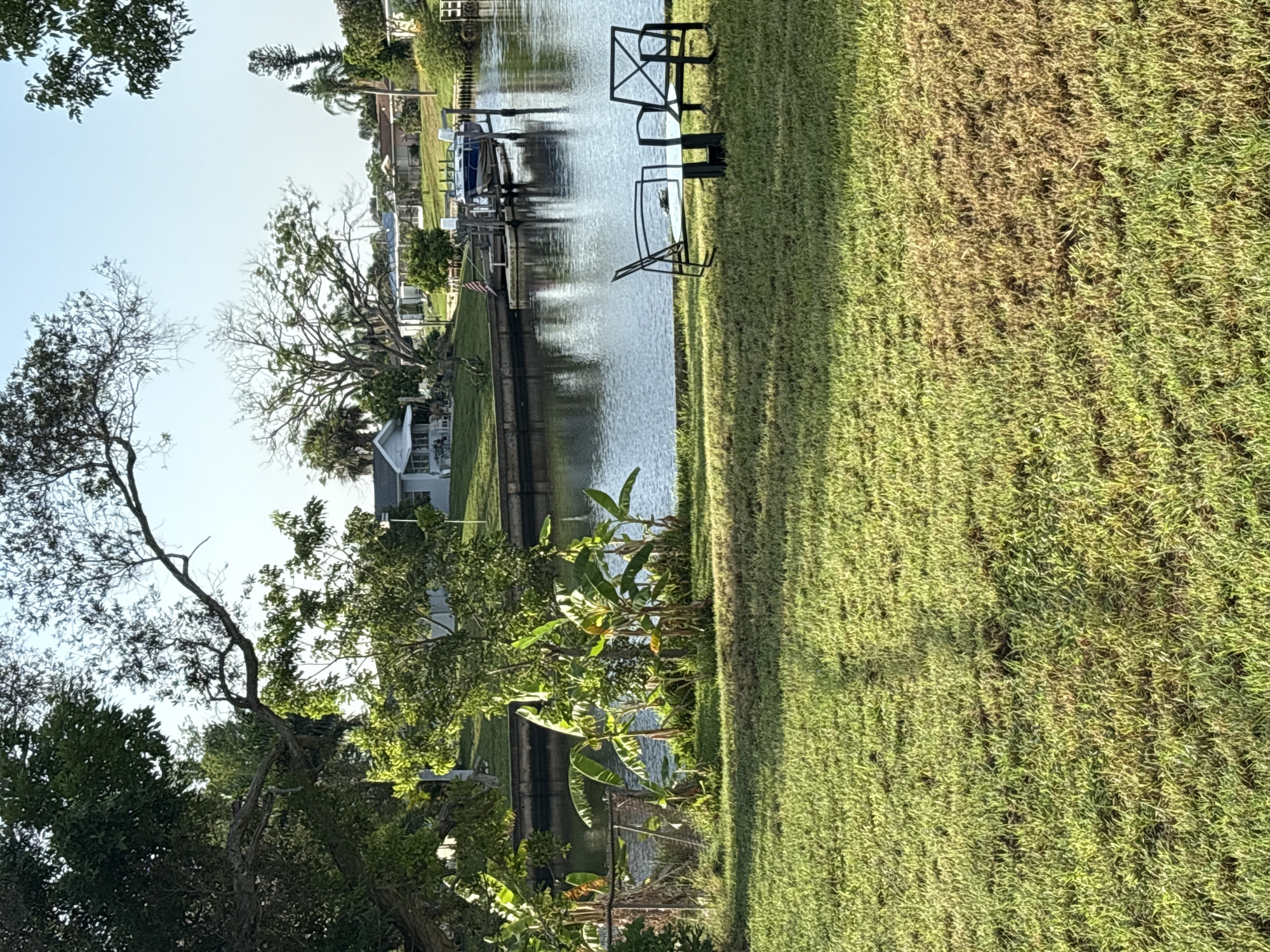 The image shows a serene waterside scene. In the foreground, a well-maintained green lawn leads to a body of water. Two black metal chairs and a glass-topped table are set on the grass, suggesting a place for relaxation. The water, likely a canal or bayou, is calm and reflects the bright blue sky. On the opposite bank of the water, several houses are visible, some with docks and boats. One boat, a blue and white vessel, is moored at a dock. Lush green trees and vegetation surround the houses, creating a peaceful, residential atmosphere. The overall impression is one of tranquility and comfortable living in a waterfront community.