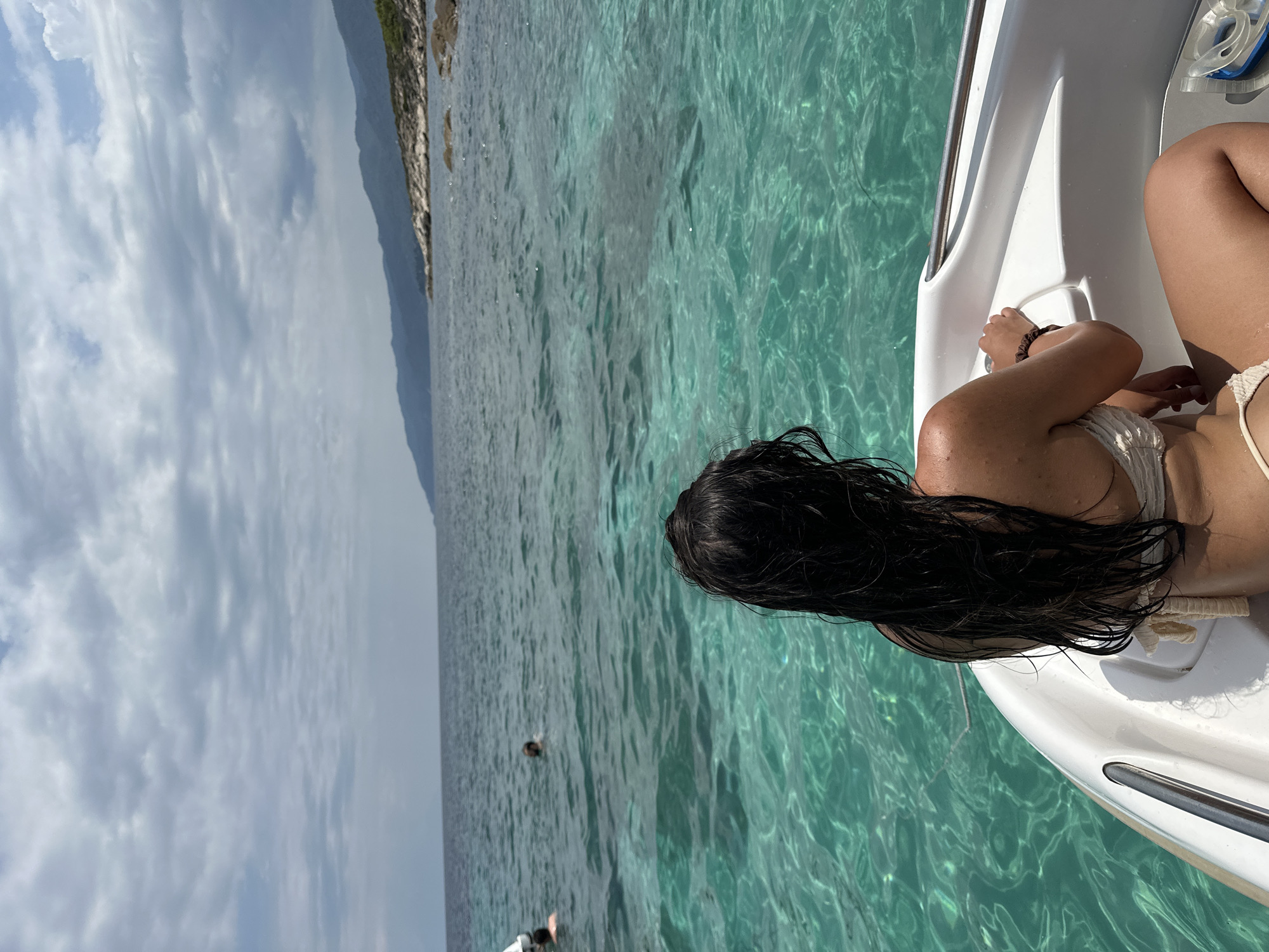 In a medium shot, a woman with dark hair pulled back sits in the front of a white boat, facing away from the camera, looking out at the ocean. She is wearing a light-colored bikini. The boat is in clear, turquoise water. In the background, two other people are visible swimming in the water. The coastline, with green hills and rocky shores, is visible in the distance under a cloudy sky.