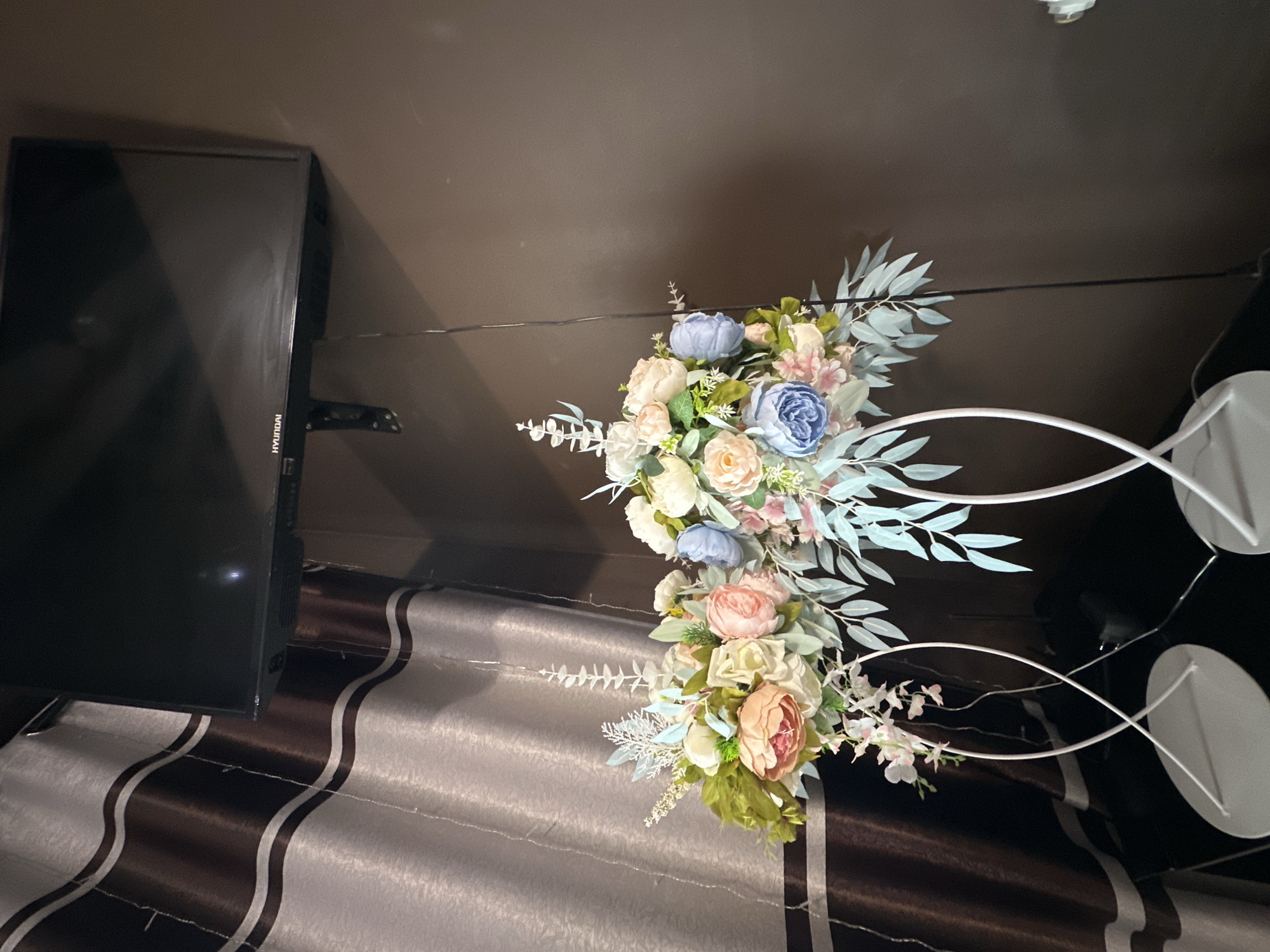 The image shows a room with a television on the left and a floral arrangement in the center. The television is black and has a "HYUNDAI" label on the side. The floral arrangement consists of roses and other flowers in shades of blue, pink, white, and peach, with some greenery. The flowers are arranged on a white metal stand. The background is a dark wall and a patterned curtain.