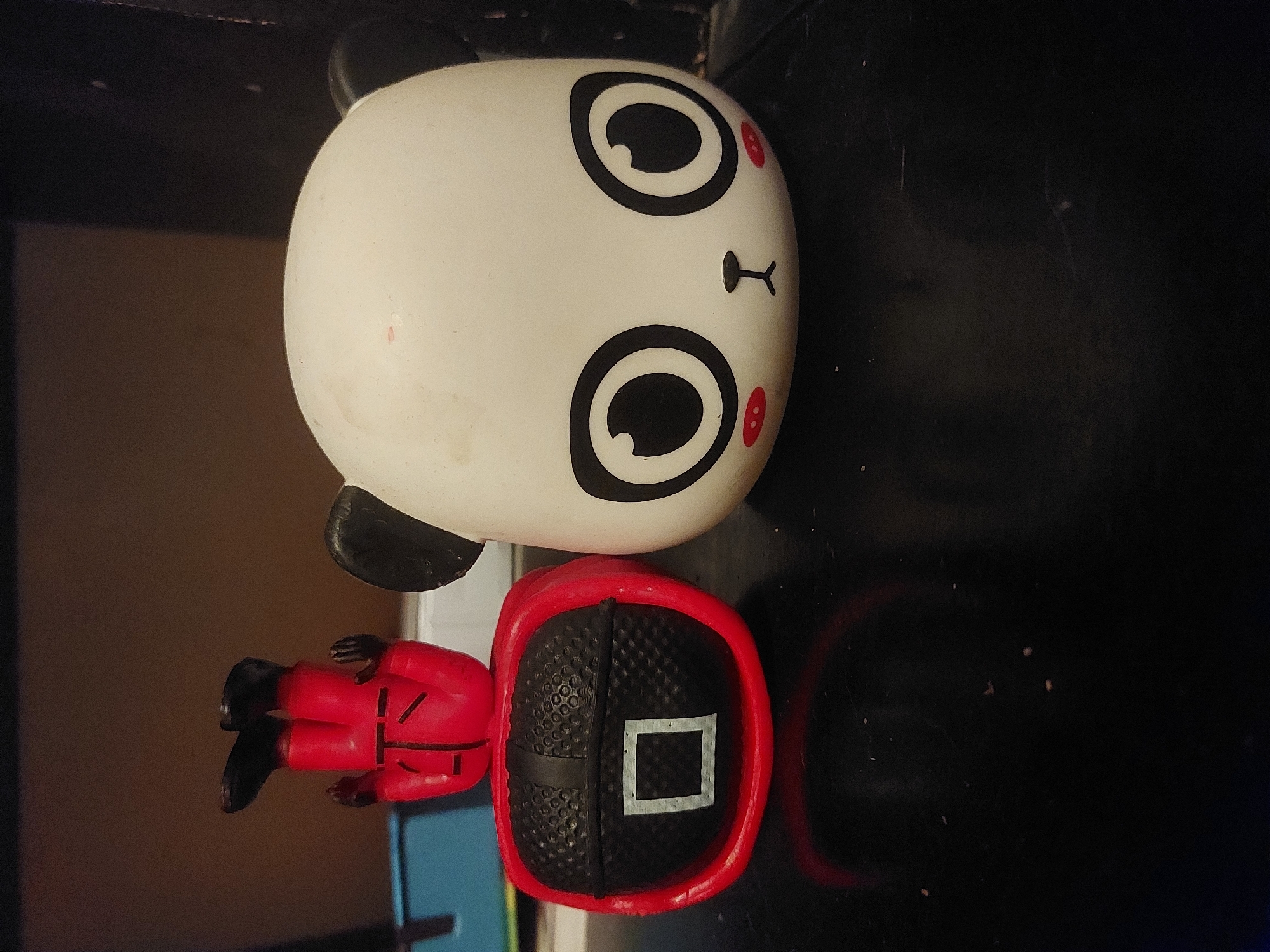 The image shows two objects lying on a dark surface. The top object is a white, rounded figure resembling a cartoon panda head with black eyes and ears, and red accents. Below it is a red figure of a Squid Game character with a black square on its head. There is nothing in the image that indicates the city of Tsarevo, Bulgaria.